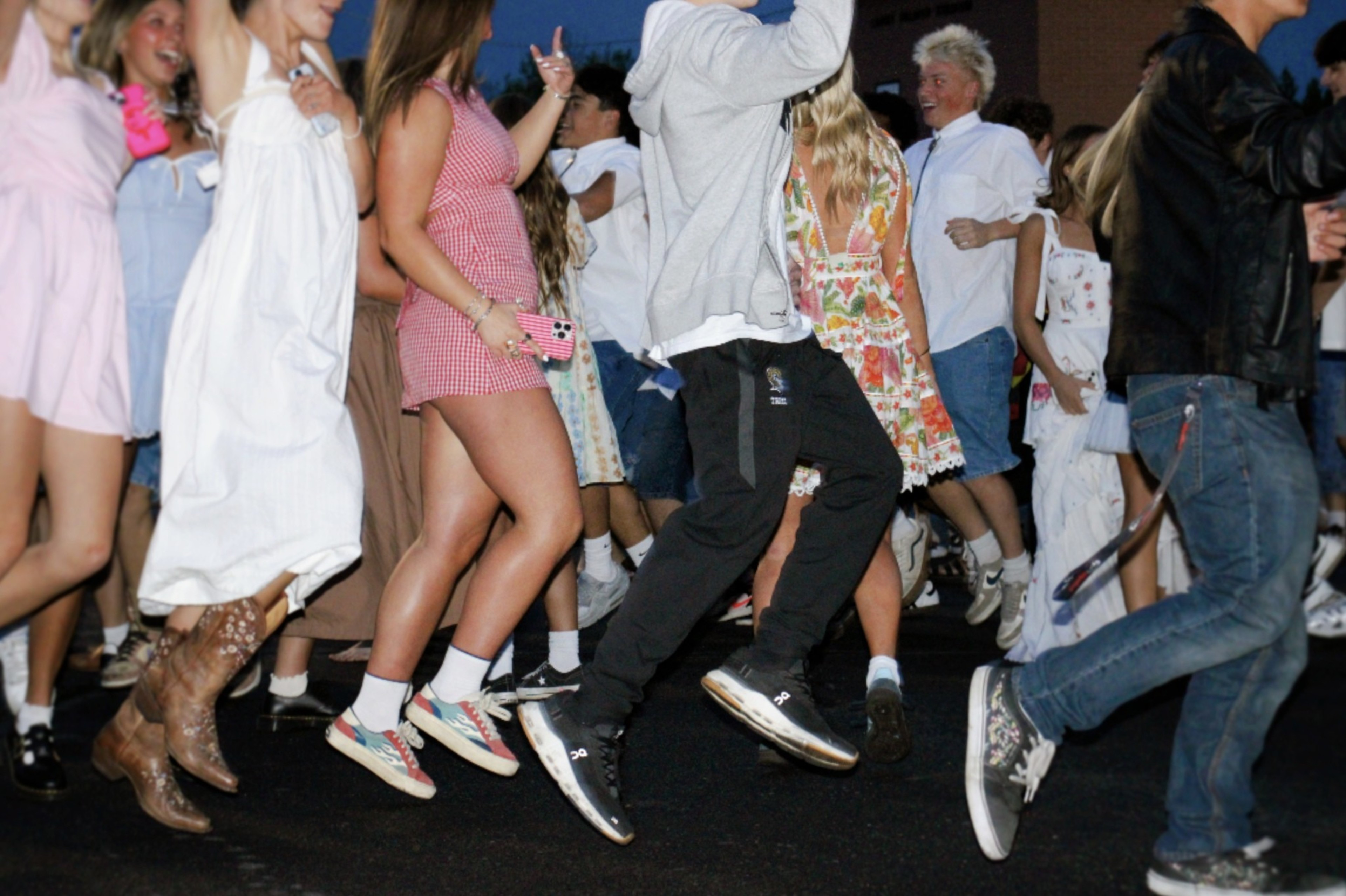 A group of people are energetically dancing outside. They're dressed in casual and summer attire, with some in dresses and sneakers, creating a lively atmosphere.