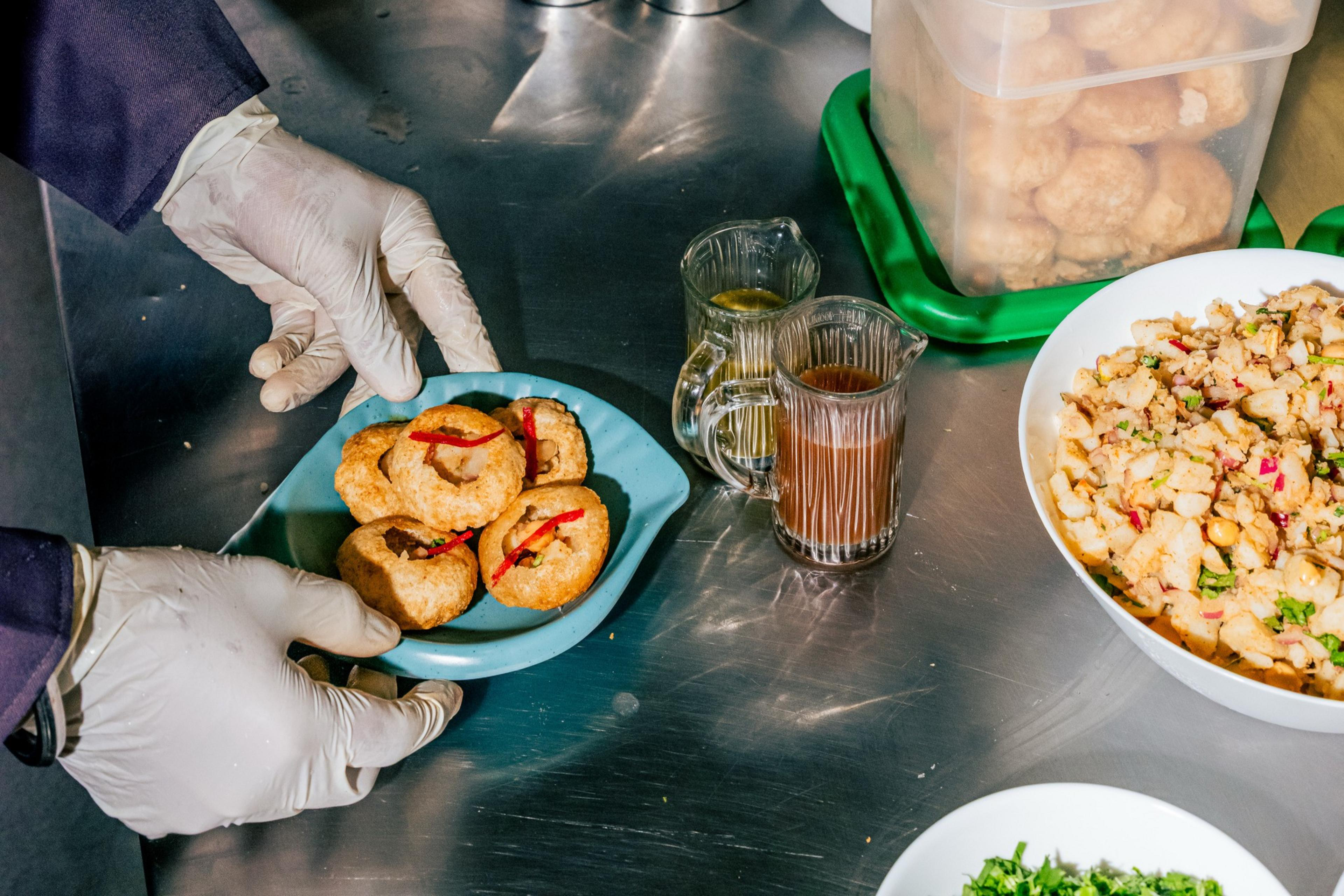 Hands wearing white gloves hold a blue dish with five fried snacks garnished with red chili slices on a metal surface next to bowls of chopped ingredients and sauces.