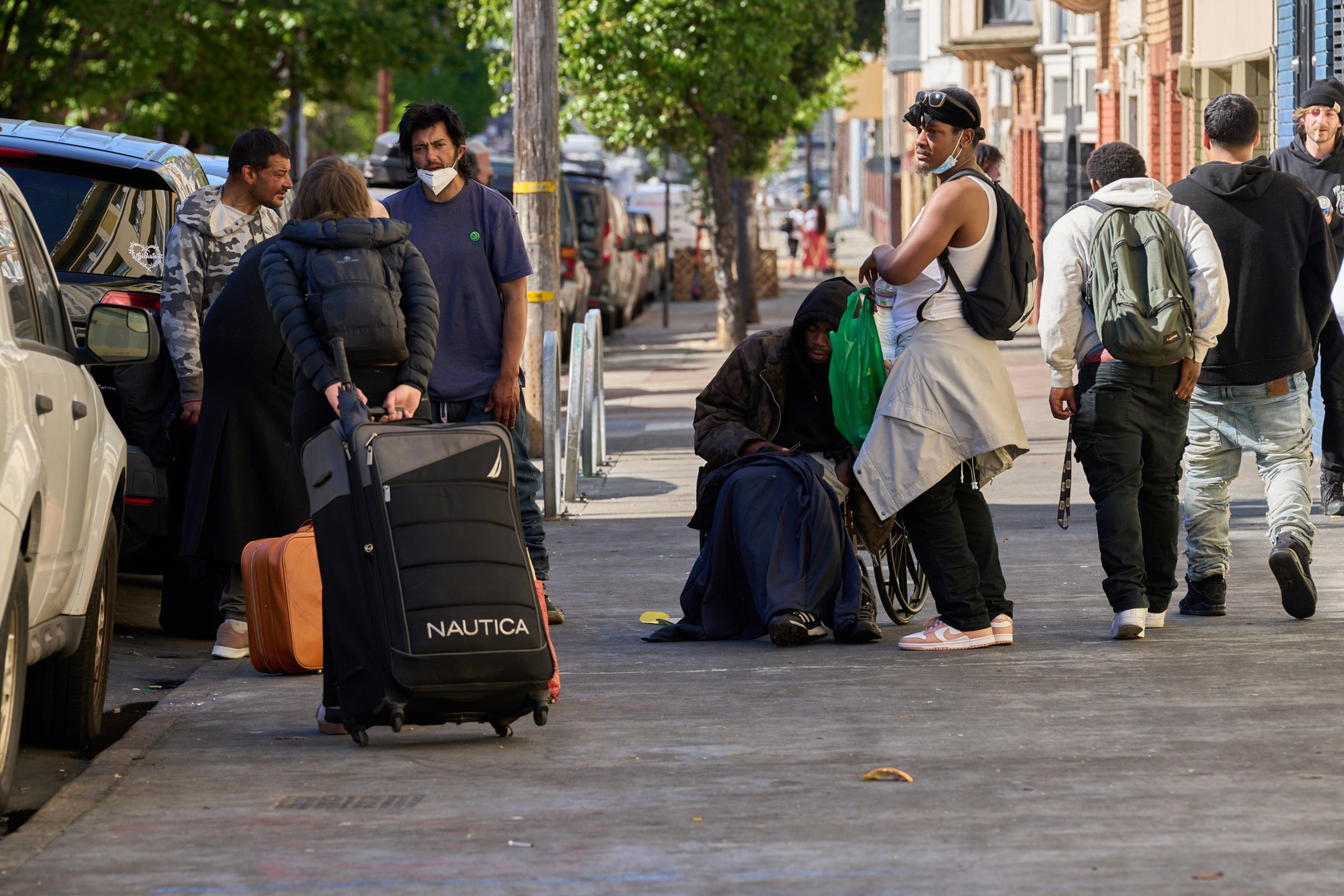 People are gathered on a sidewalk beside parked cars, some with luggage, others engaging in conversation. Trees line the street in the background.