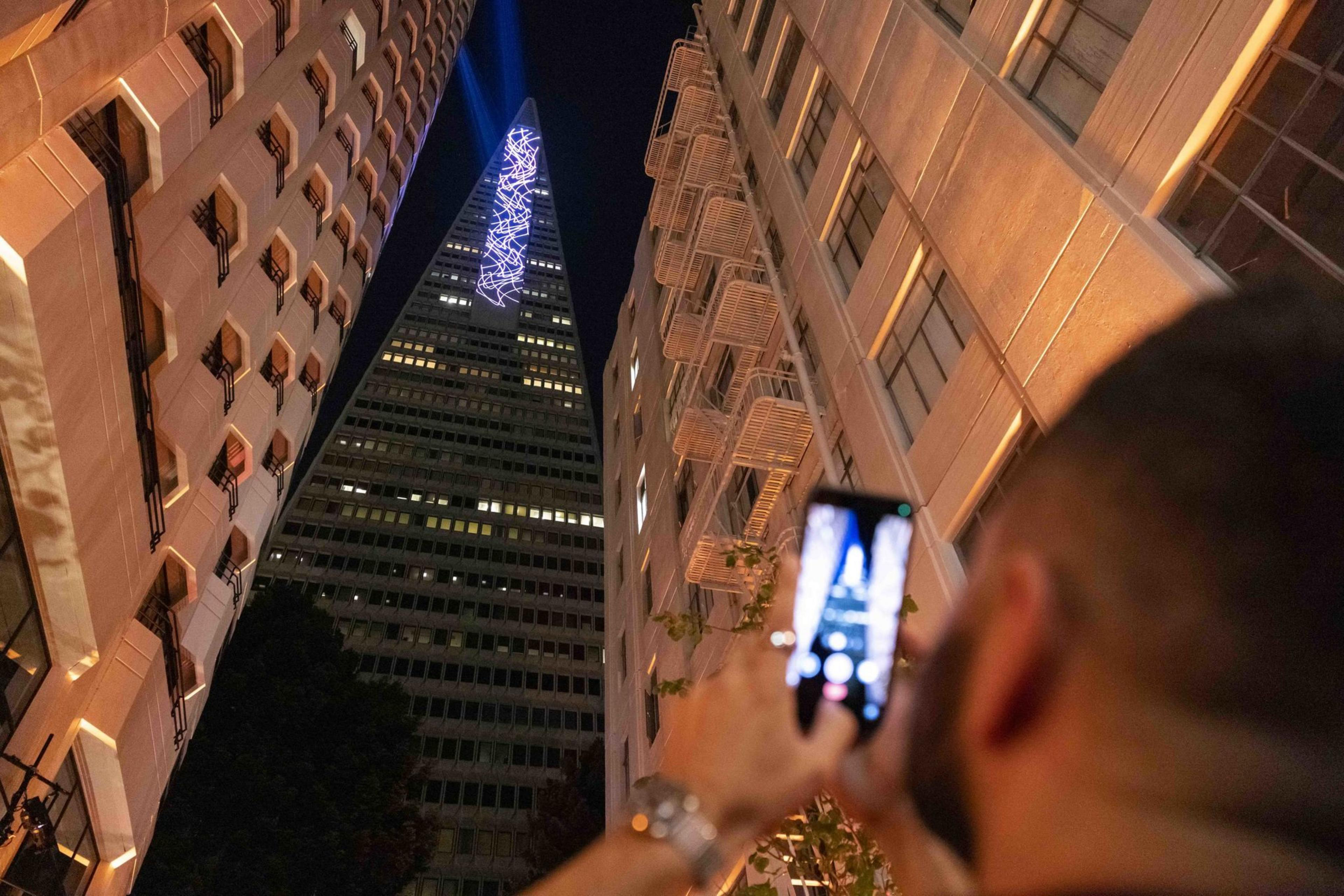 A person is photographing a tall, pointed skyscraper with colorful lights at night, flanked by two other buildings.