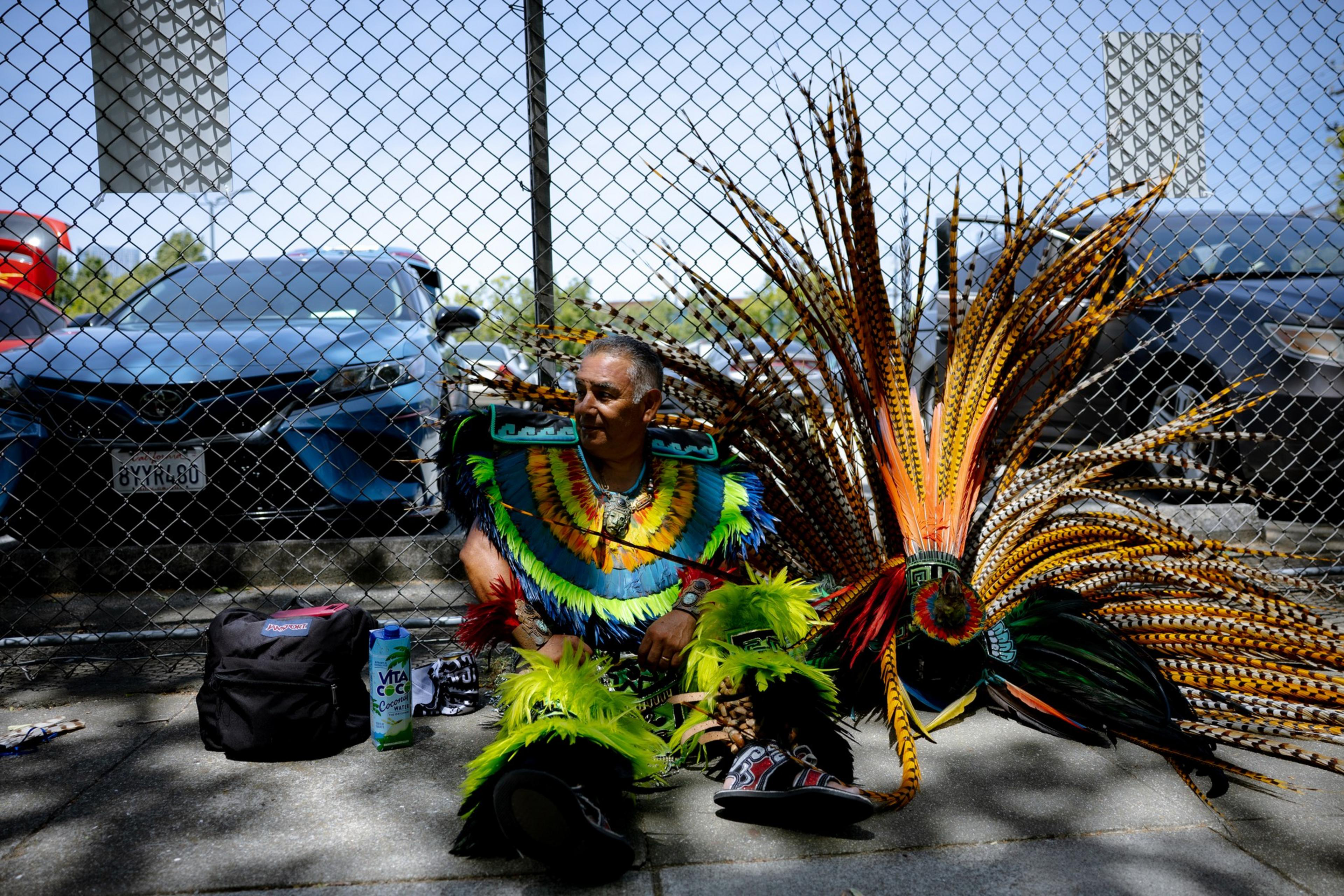 The image shows a man sitting against a chain-link fence, wearing vibrant, colorful traditional attire with feathered decorations. Cars and bags are nearby.