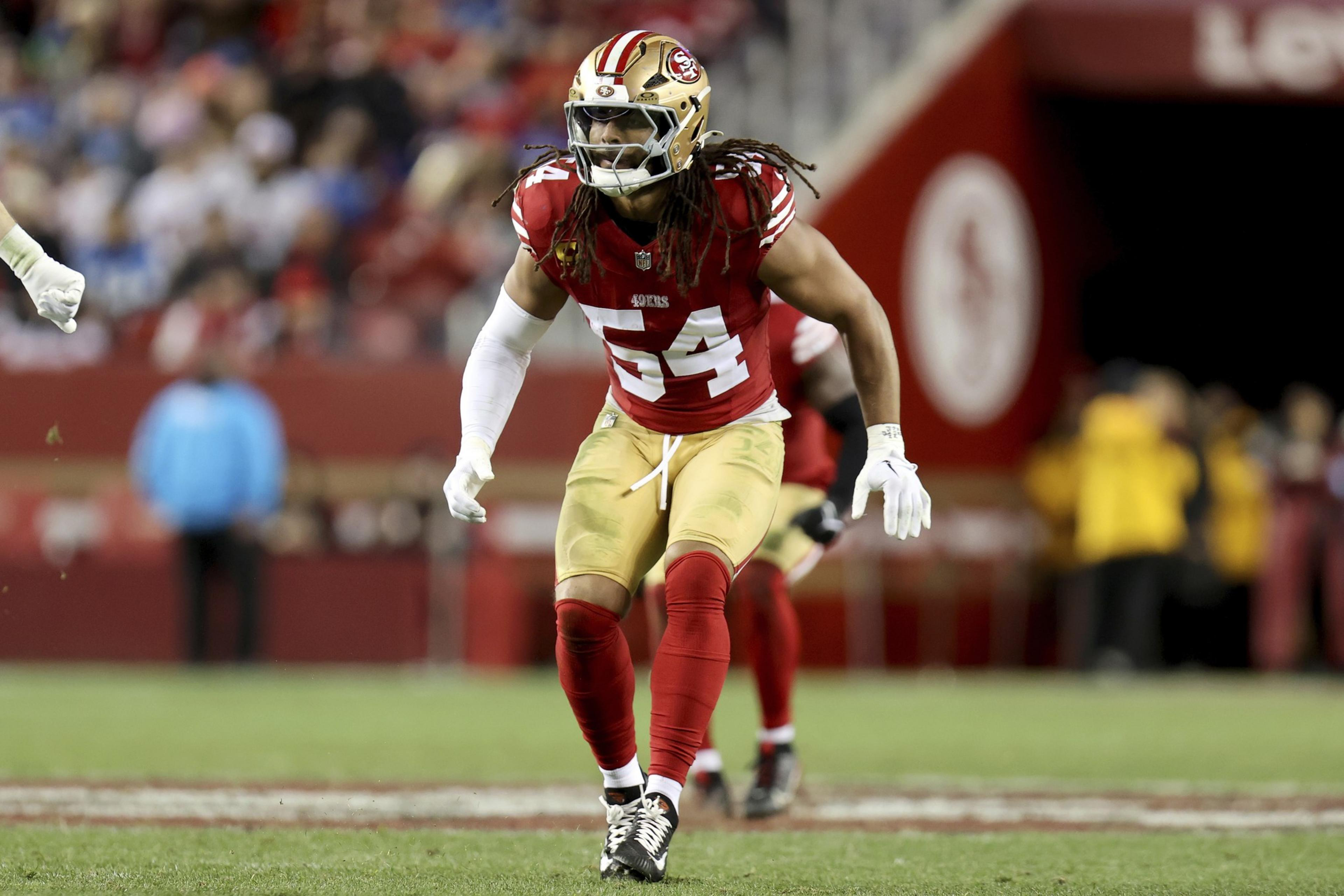 A football player in a red and gold uniform is mid-action on the field, wearing a helmet and visor. The background is a blurred stadium.