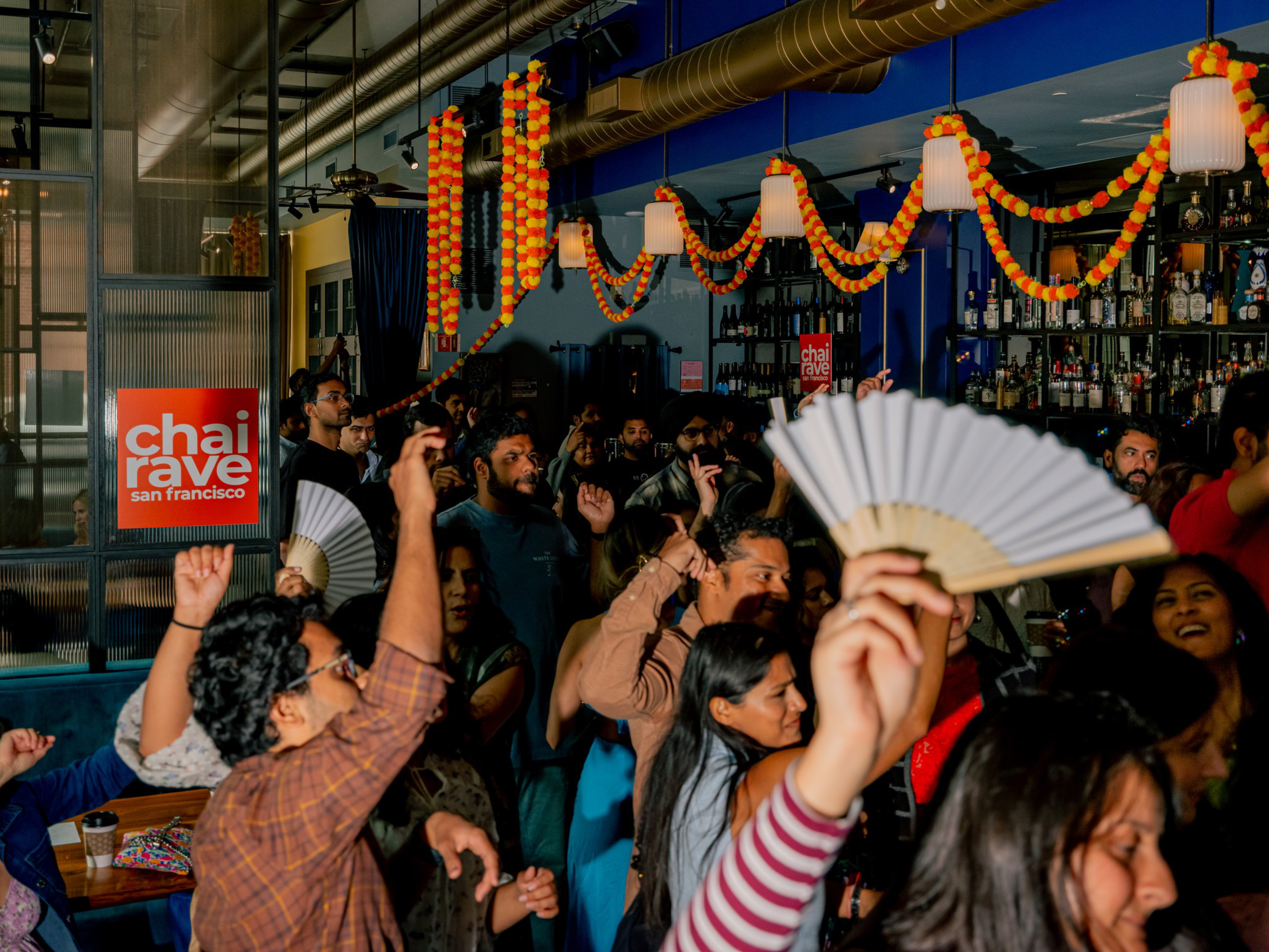 A lively crowd dances and waves fans inside a bar decorated with orange and yellow garlands at Chai Rave San Francisco.