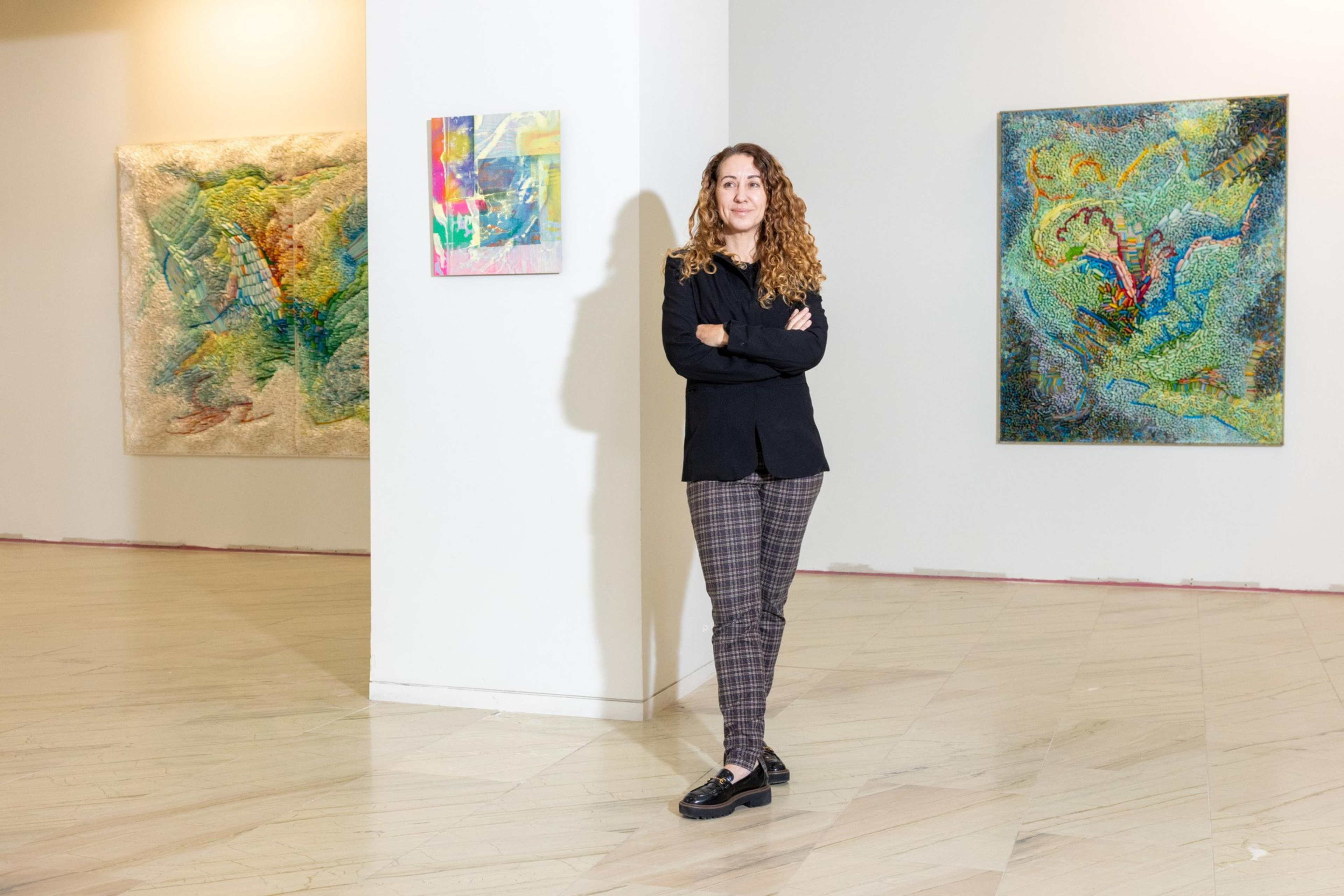 A woman with curly hair stands with arms crossed in a gallery, surrounded by vibrant, textured abstract paintings on white walls.