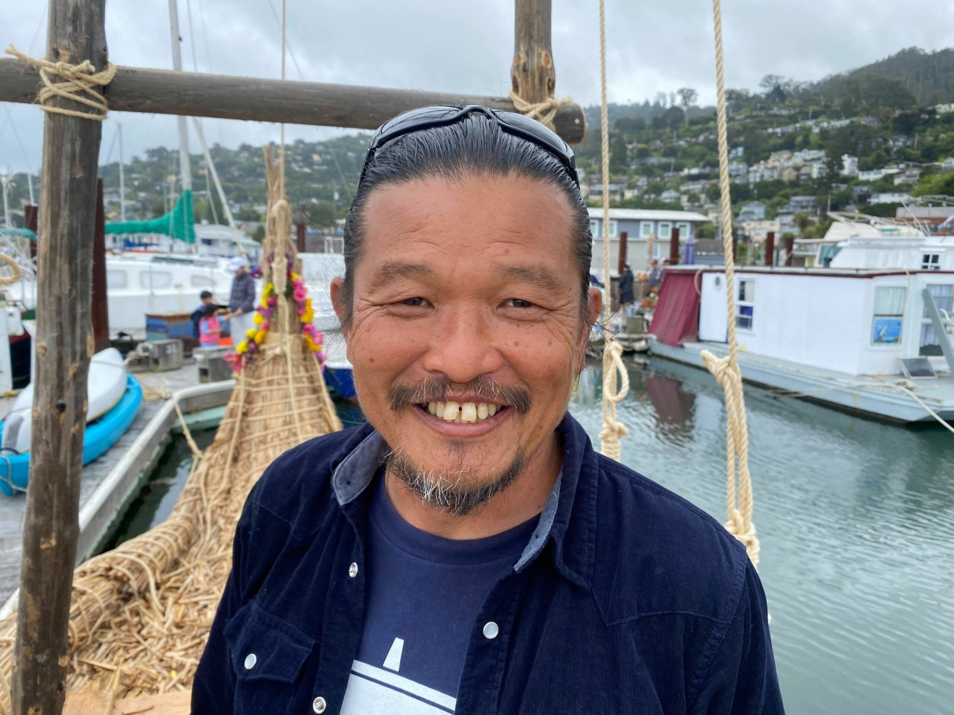 A smiling man with a beard and mustache stands in front of a woven reed boat at a marina with boats and hills in the background.