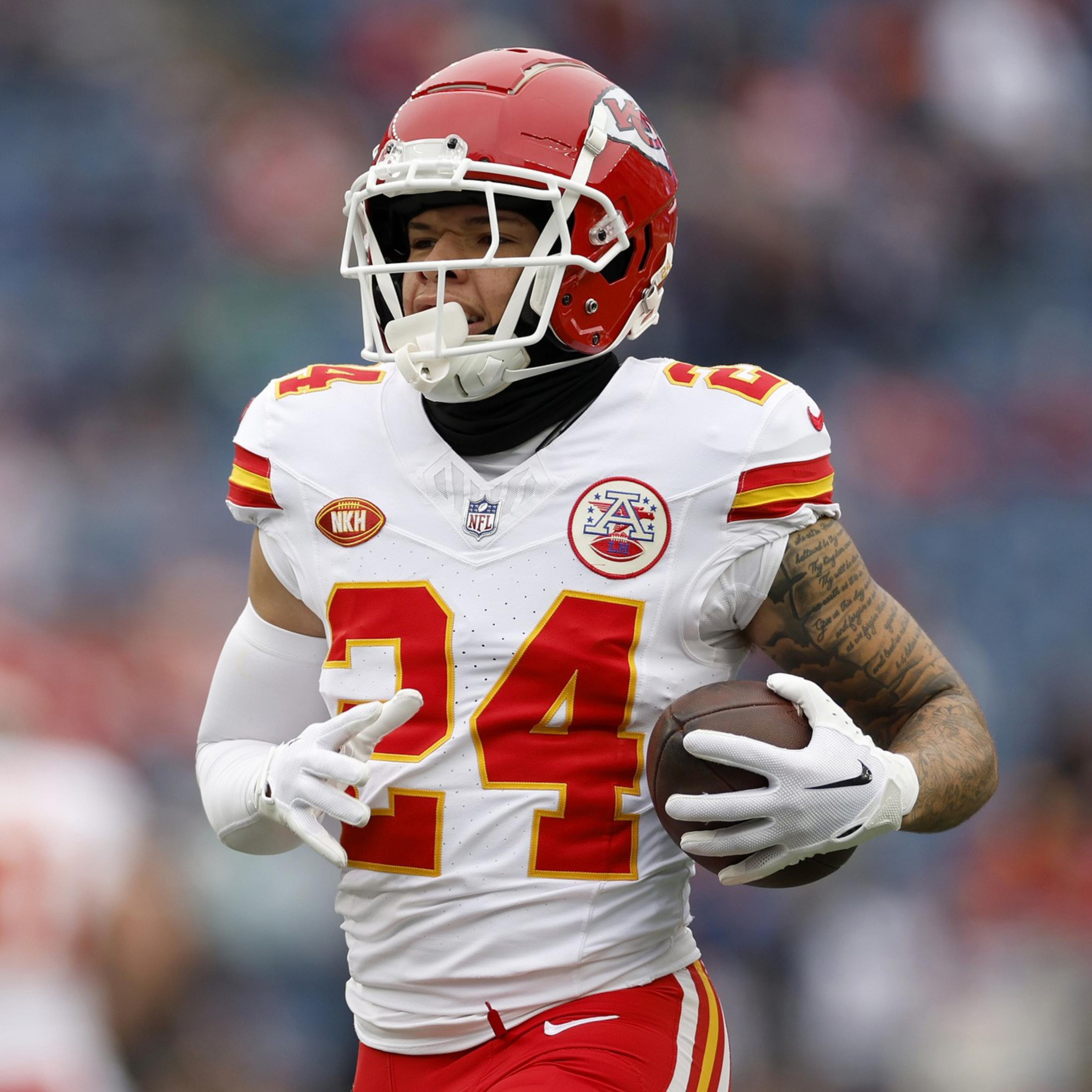 A football player in a red helmet and white jersey with number 24 is holding a football and running on the field.