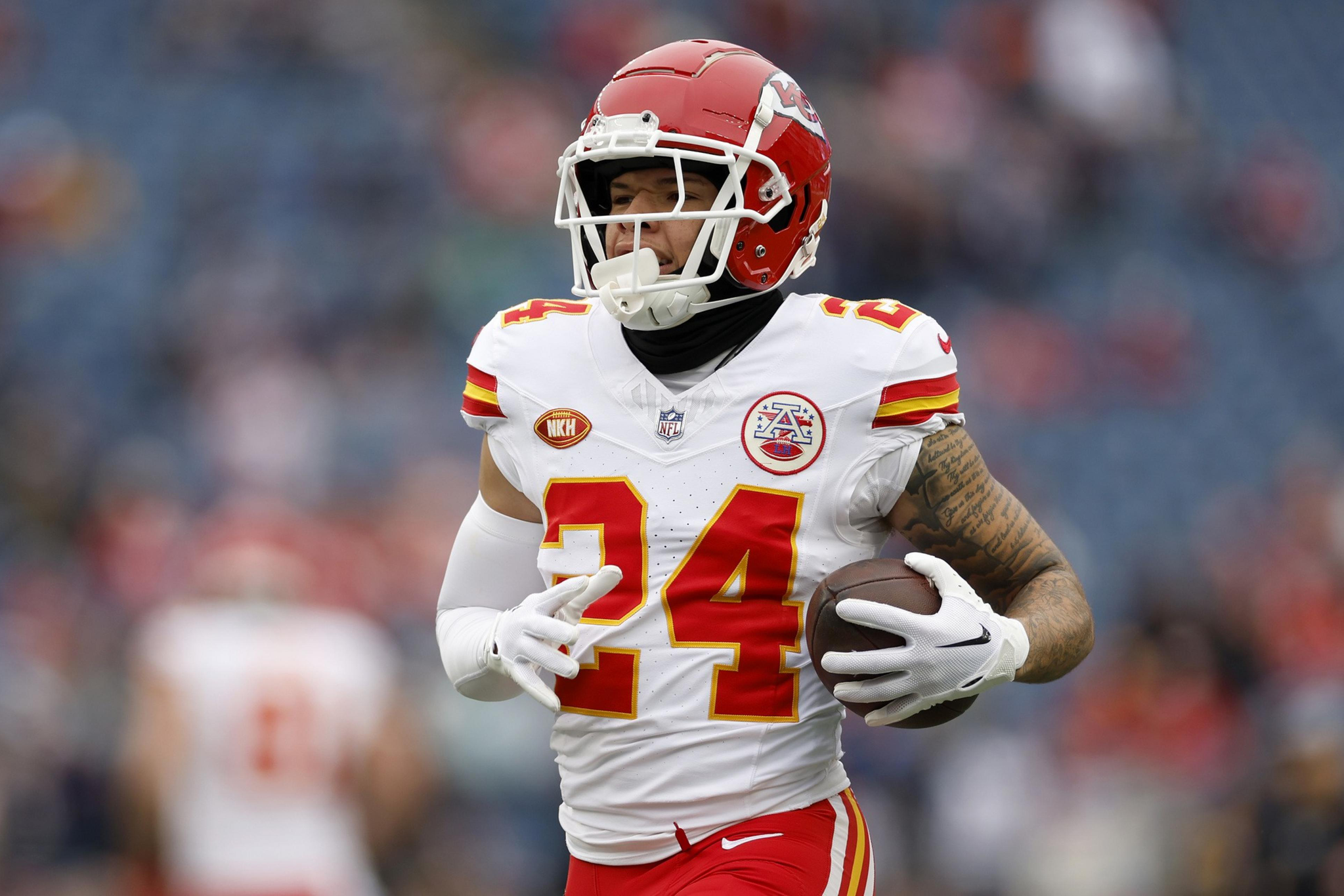 A football player in a red helmet and white jersey with number 24 is holding a football and running on the field.