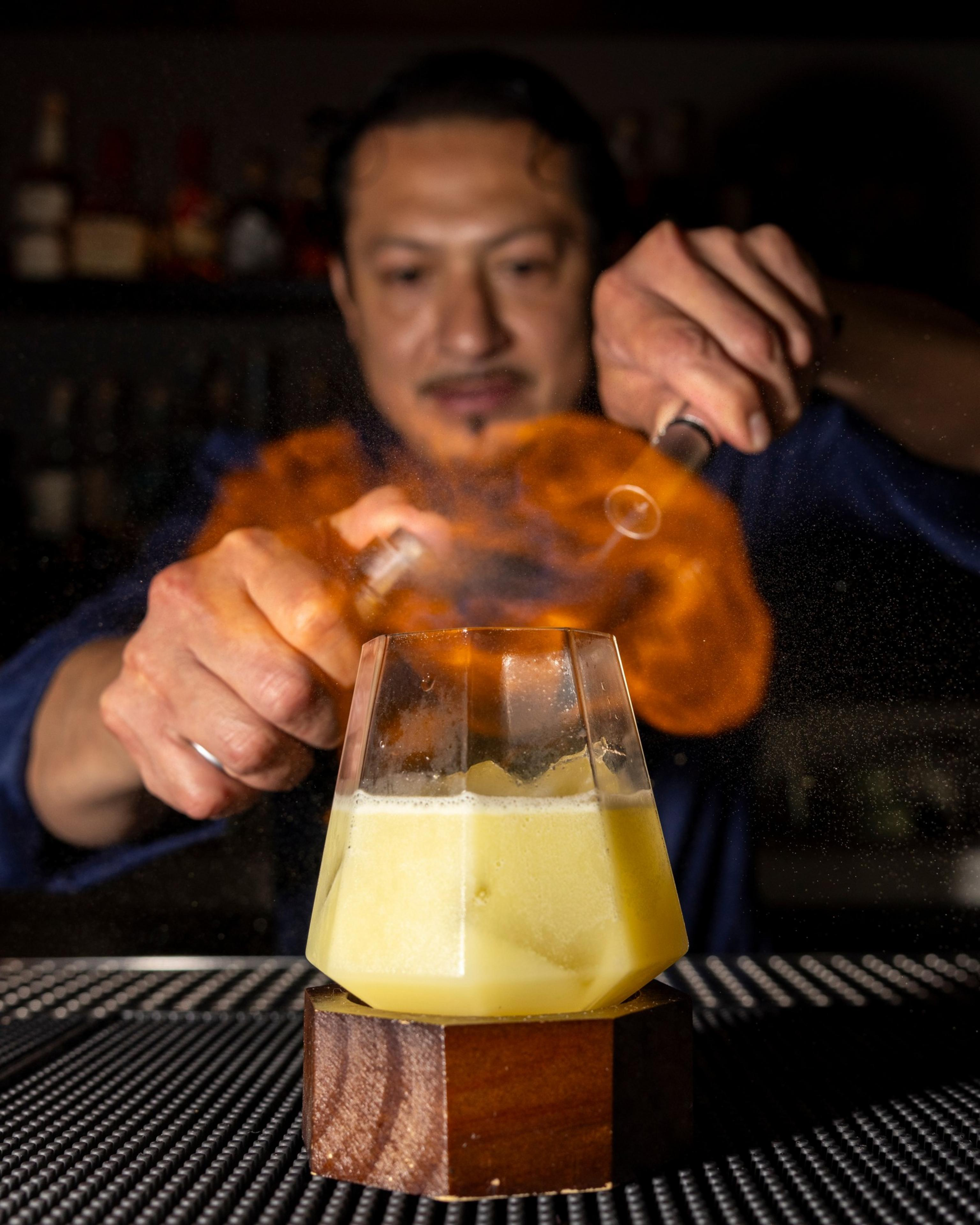 A bartender uses a flame to garnish a yellow drink in a geometric glass, with a burst of fire surrounding the drink on a dimly lit bar.