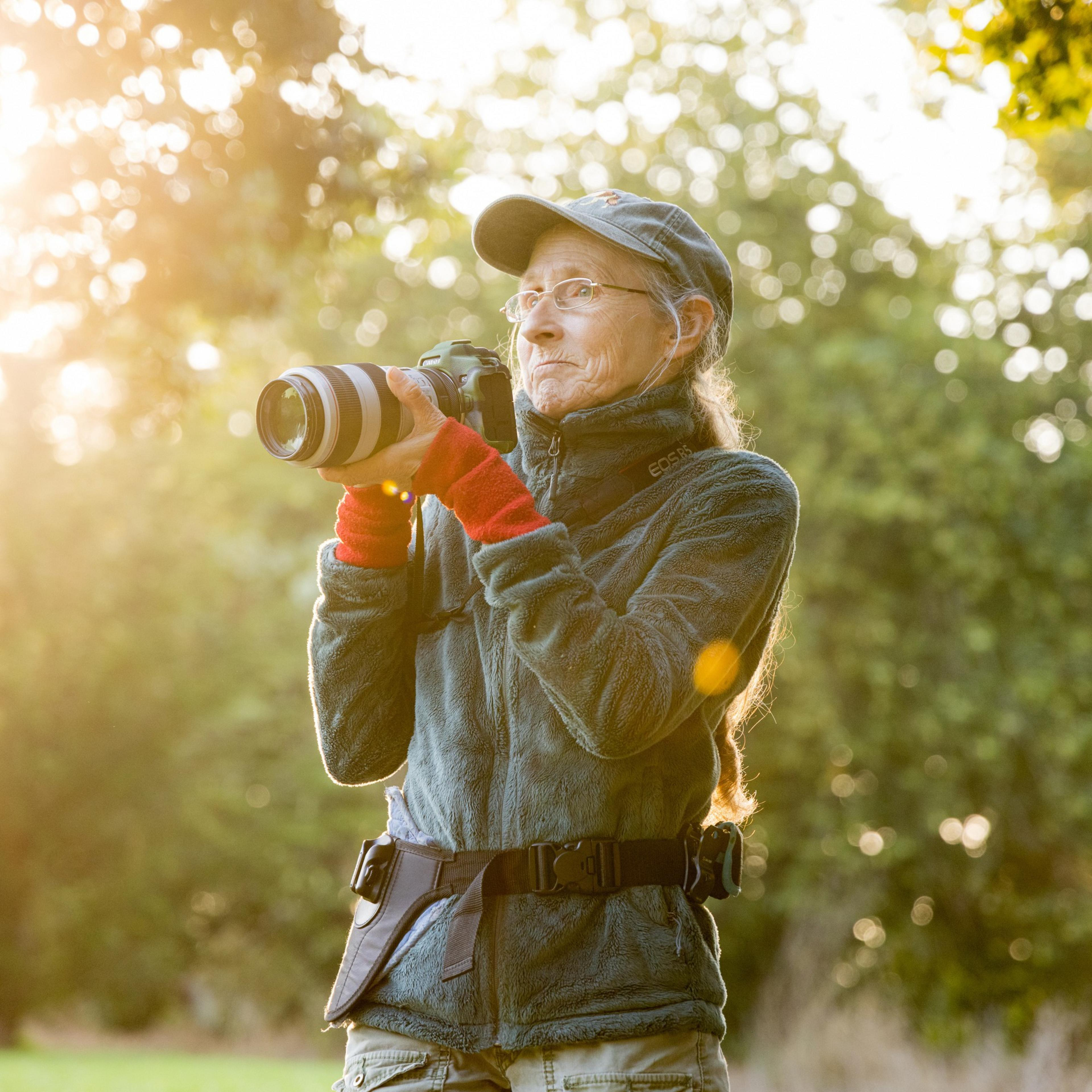 An individual with glasses and a cap stands outdoors, holding a camera with a long lens. They are wearing warm clothing and are bathed in a golden, sunlit glow.