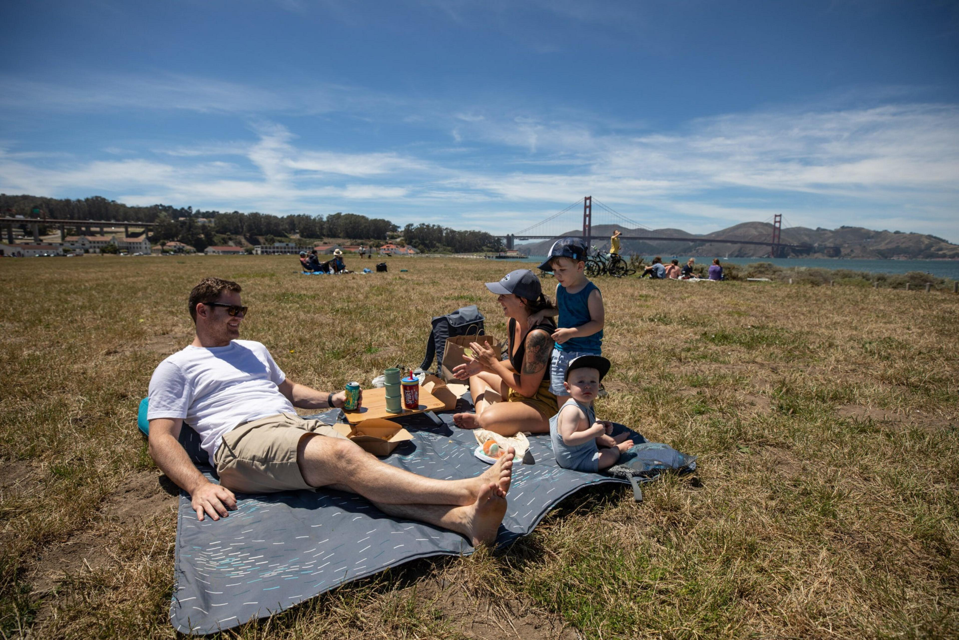 A family of four enjoys a picnic on a blanket in a grassy field with the Golden Gate Bridge and blue sky in the background.