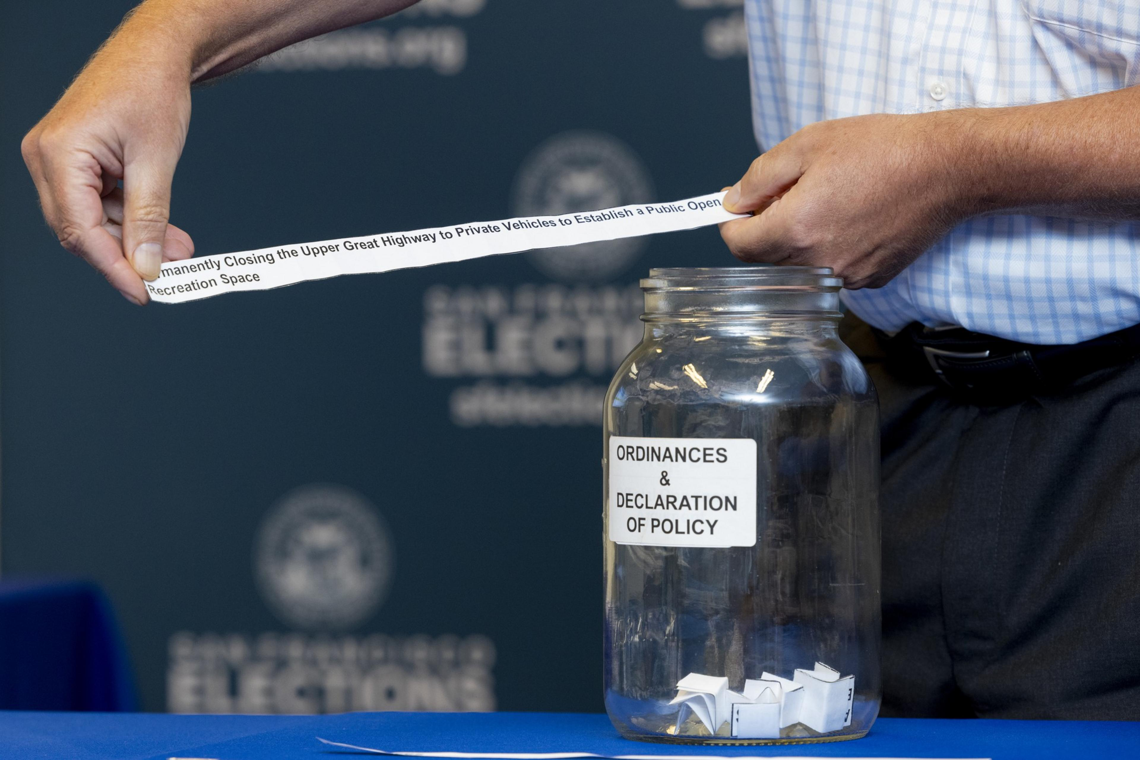 Two hands pull a strip of paper reading &quot;Permanently Closing the Upper Great Highway to Private Vehicles...&quot; from a glass jar labeled &quot;Ordinances &amp; Declaration of Policy.&quot;