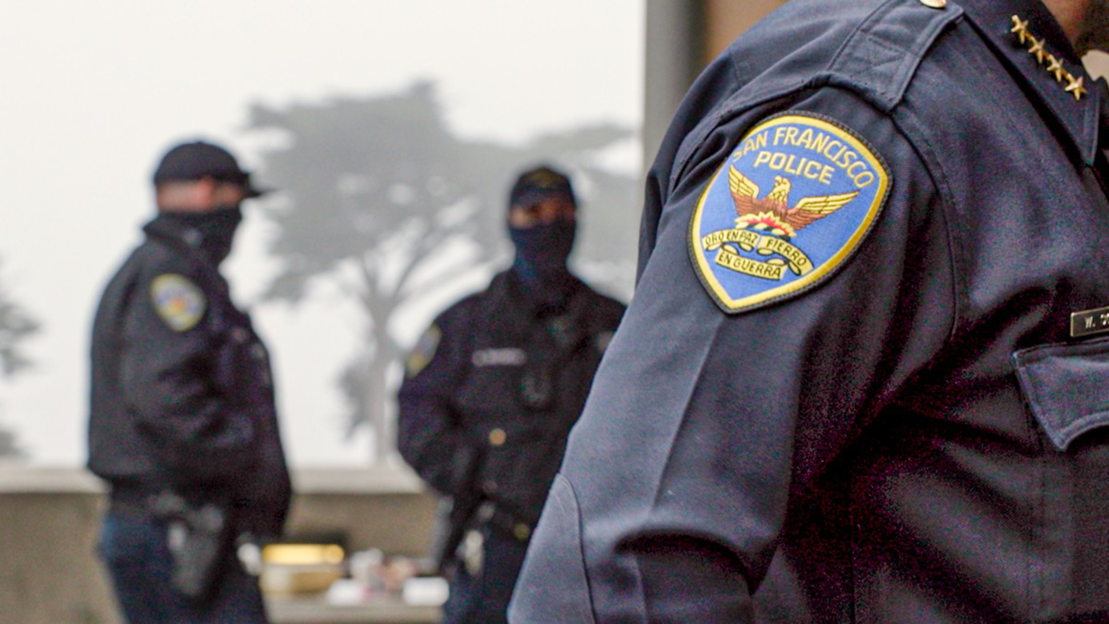 A close-up of a San Francisco Police officer's uniform patch is in the foreground, with two other masked officers standing blurred in the foggy background.