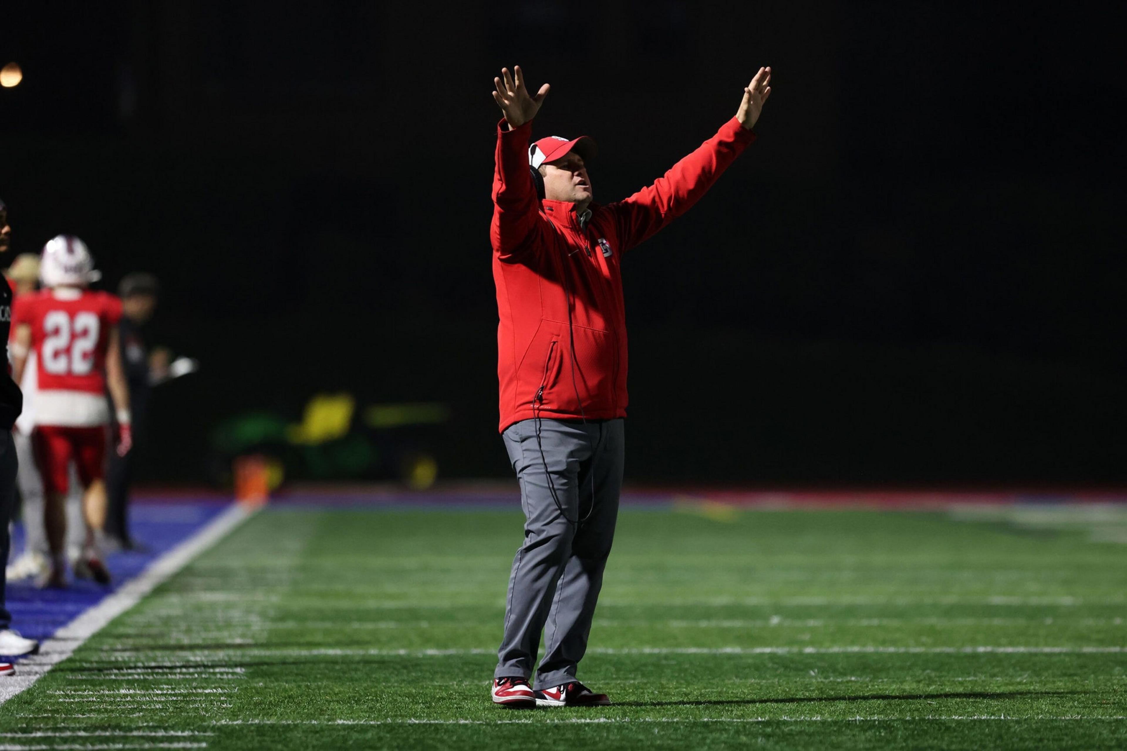 A man in a red jacket and cap stands on a football field with arms raised, while players in red and white uniforms walk away in the background.