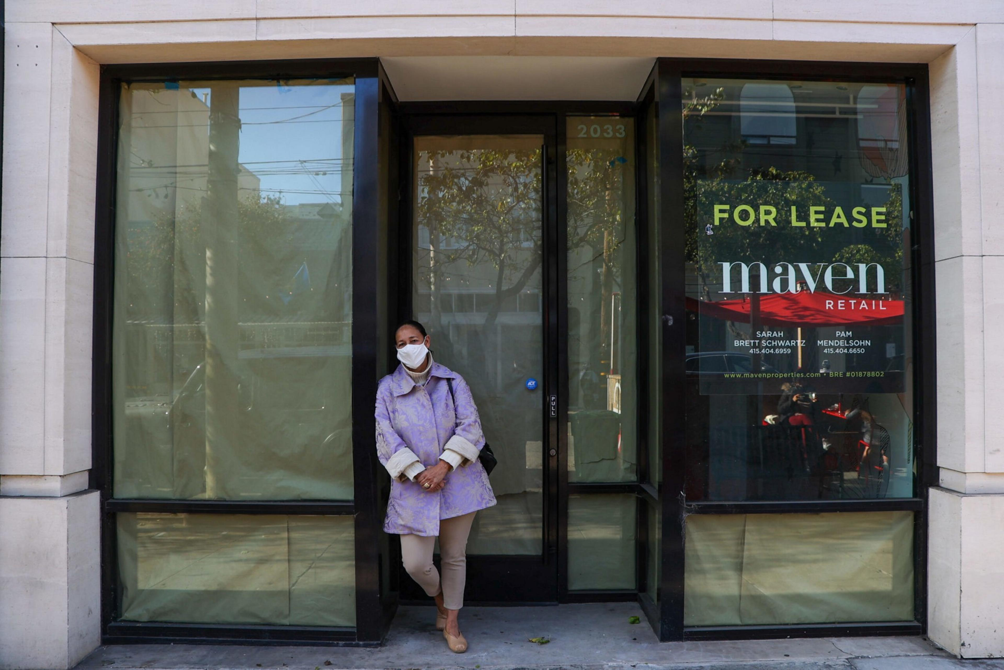 A person wearing a mask and a purple coat leans against the doorframe of an empty retail space with "For Lease" signs in the windows.