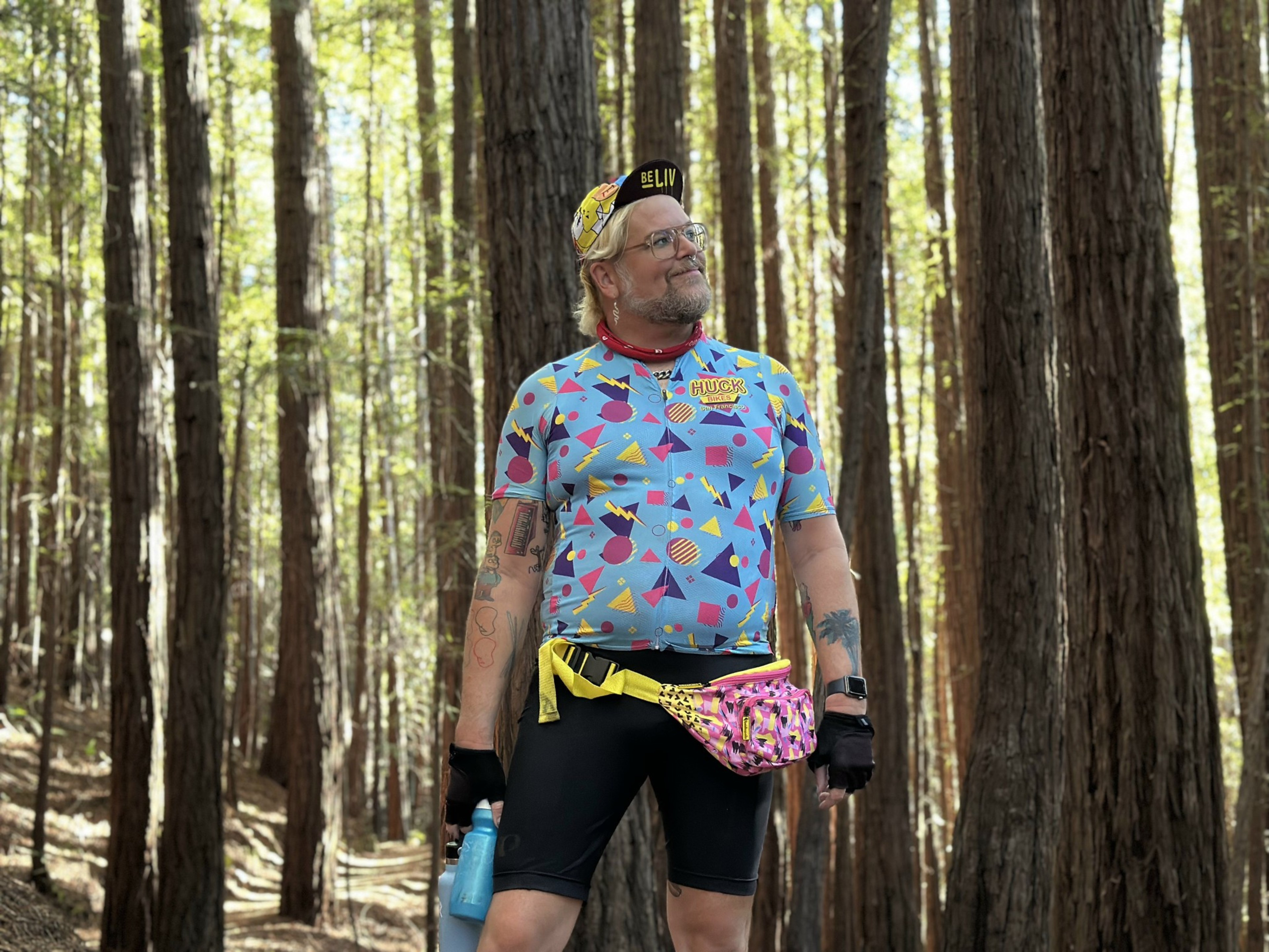 A person wearing a colorful geometric-patterned cycling jersey, black shorts, a yellow and pink fanny pack, and a cap stands in a sunlit forest holding a water bottle.
