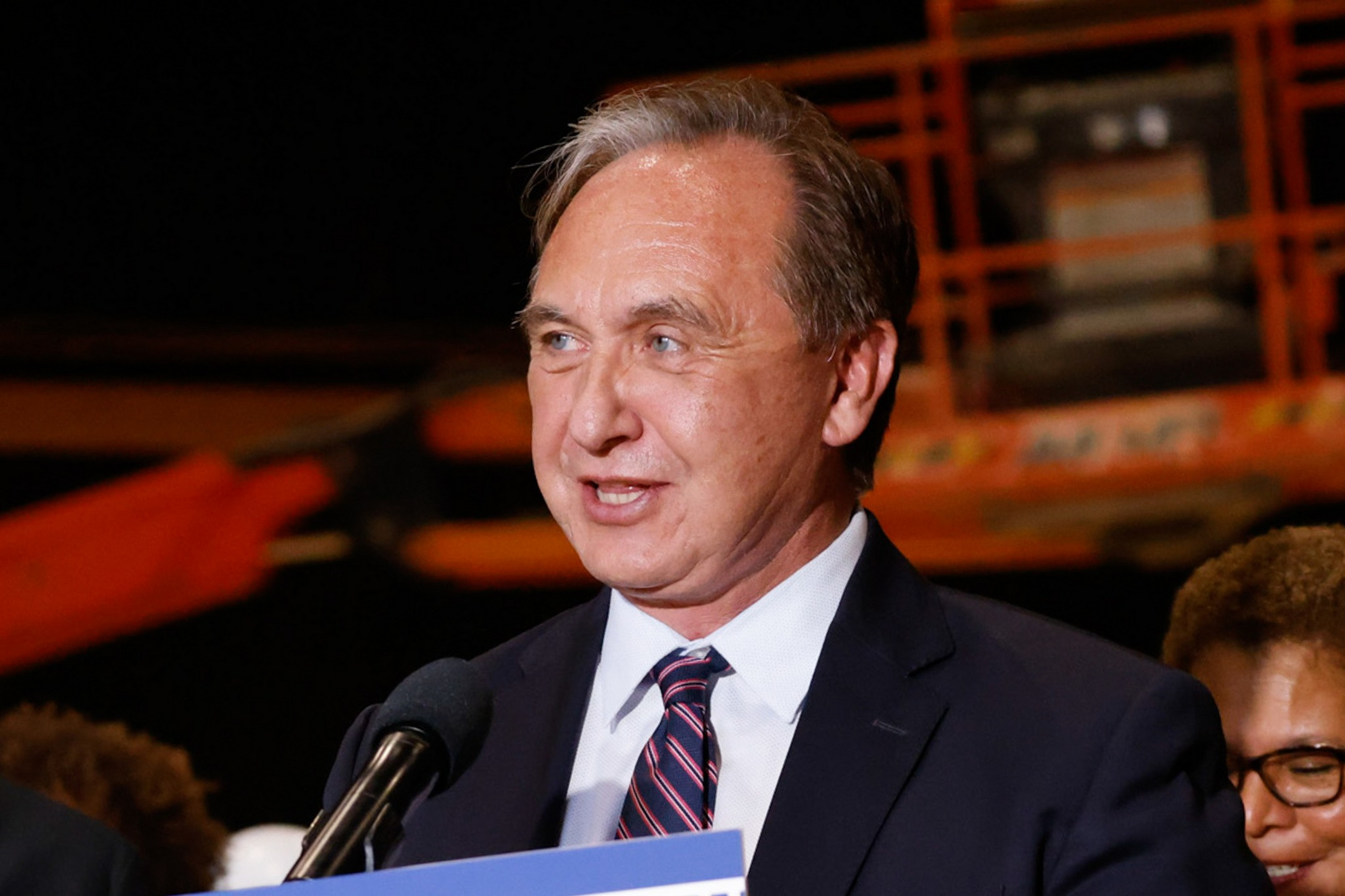 A middle-aged man in a suit and striped tie speaks into a microphone, with blurred people and orange industrial equipment in the background.