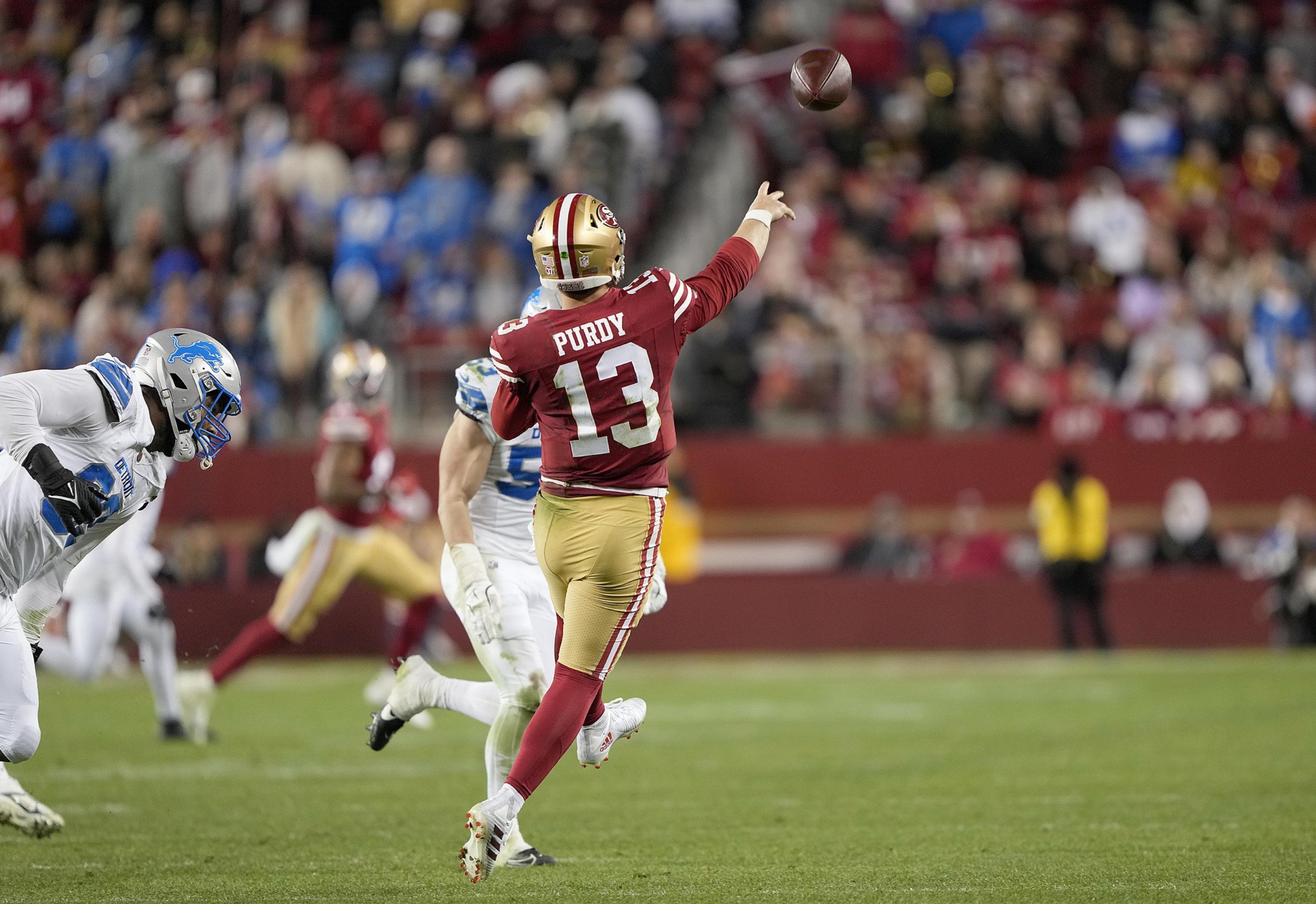 A football player in a red uniform throws a pass while evading a player in a white uniform. The crowd in the background is blurred, focusing on the action.