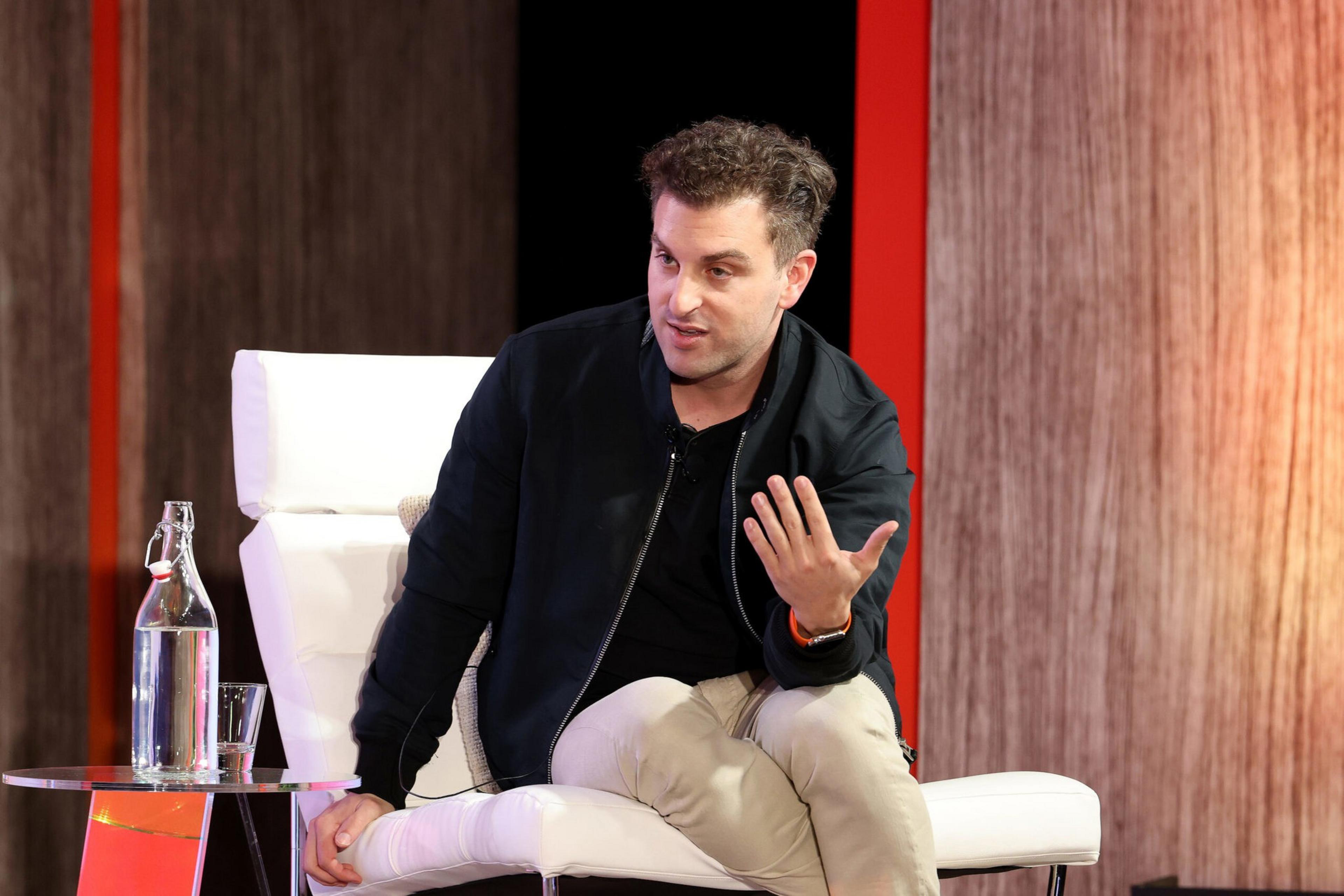 A man wearing a black jacket and beige pants sits on a white chair, gesturing with his right hand, with a glass bottle and cup on a nearby table.