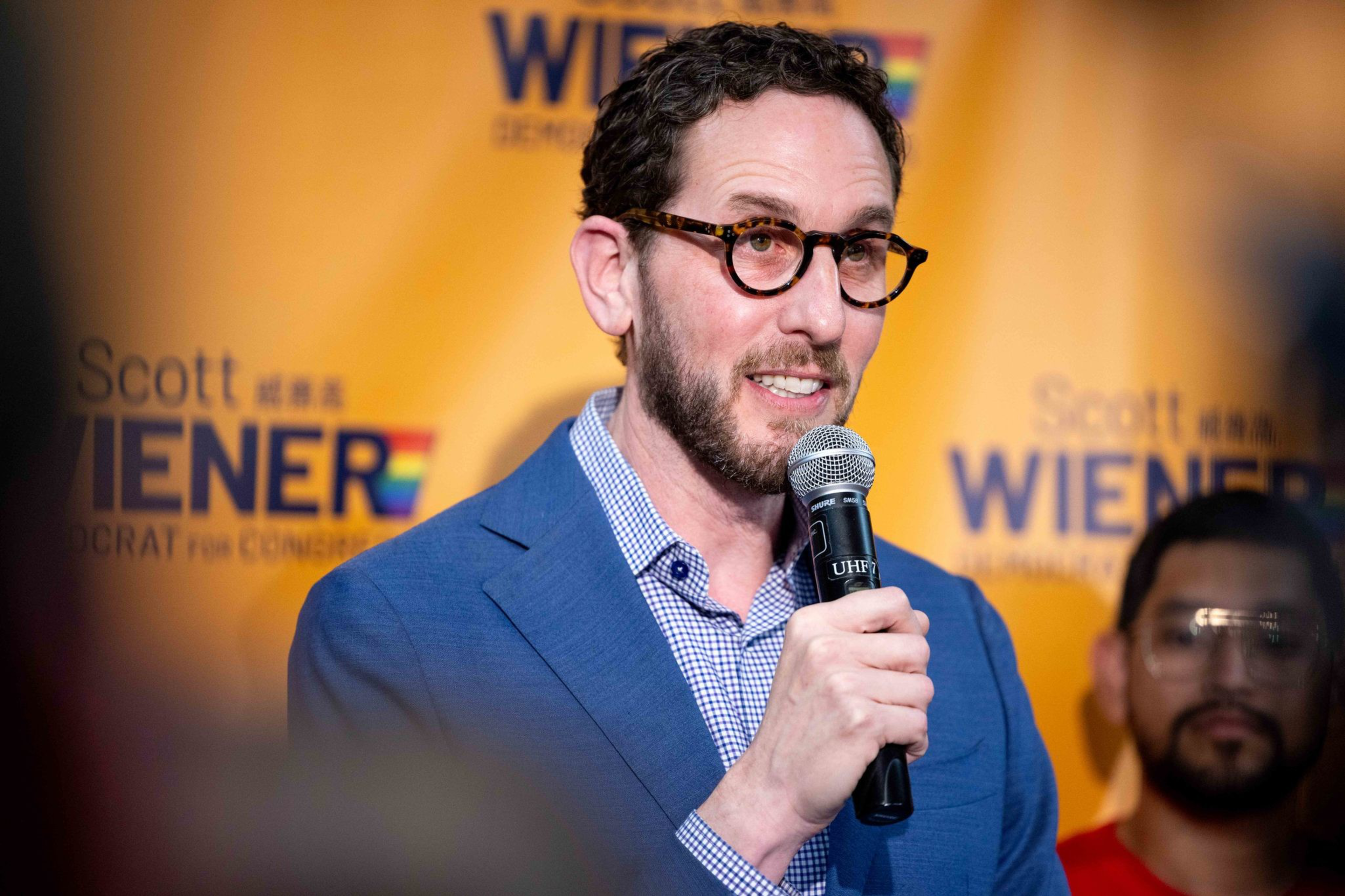 A man with curly hair, glasses, and a beard speaks into a microphone, wearing a blue blazer over a checkered shirt, with a yellow background.