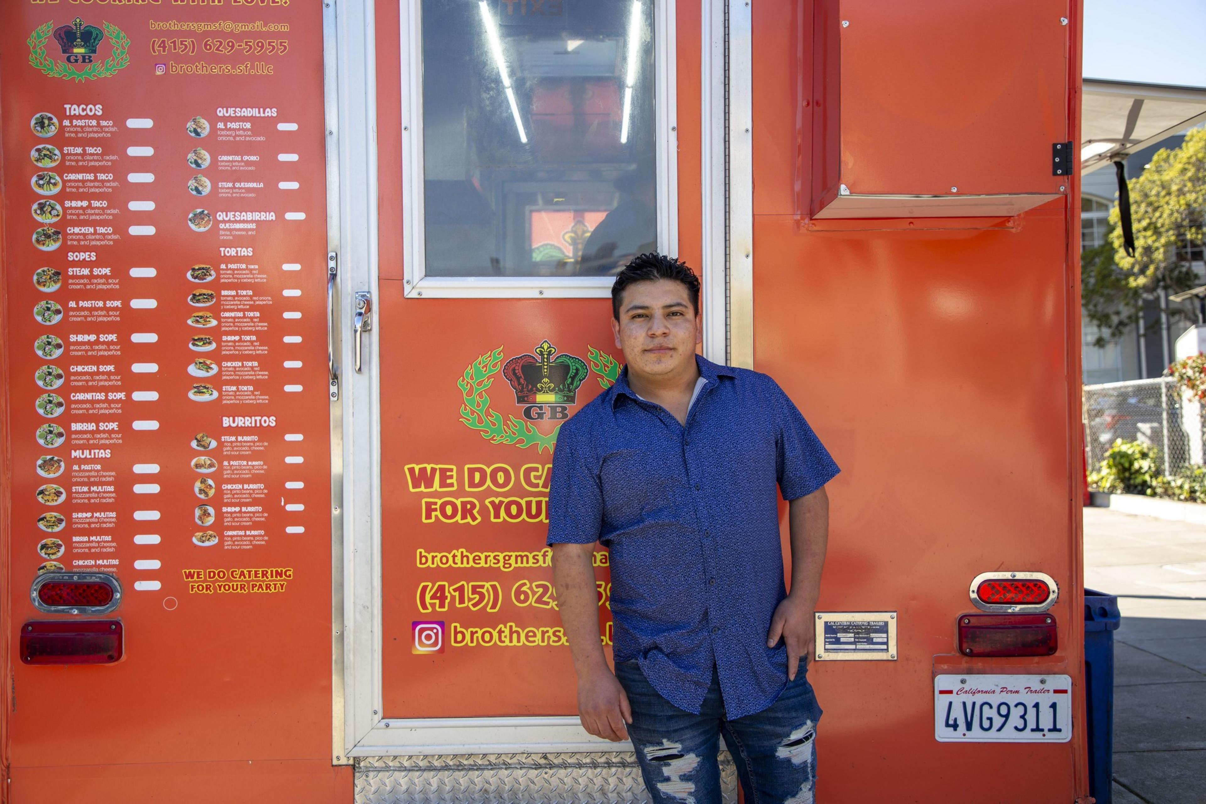 A person in a blue shirt leans against an orange food truck with a visible menu and contact details. The truck offers various Mexican dishes.