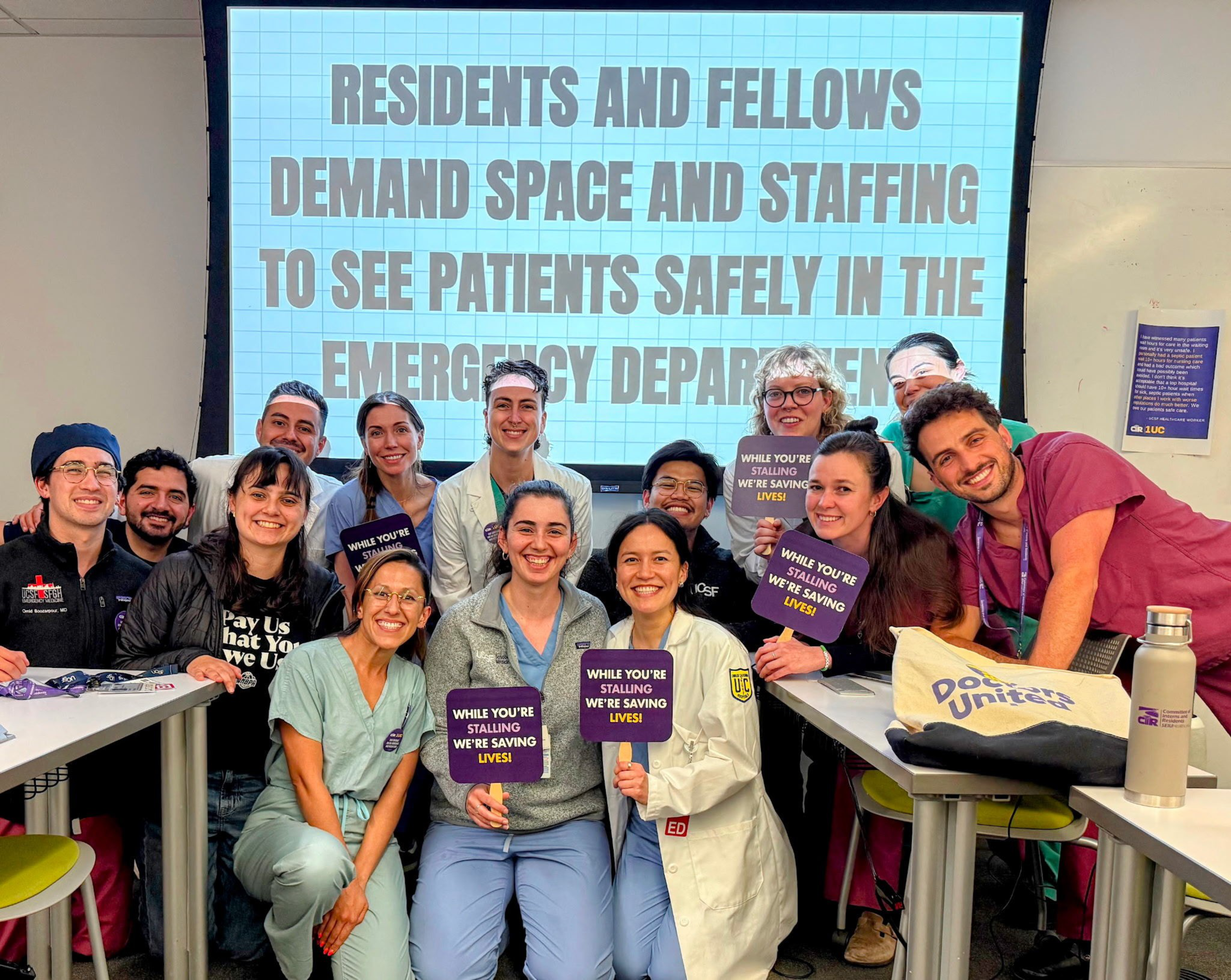 Residents and fellows hold signs saying “While you’re stalling we’re saving lives” in front of a screen demanding space and staffing in the emergency department.