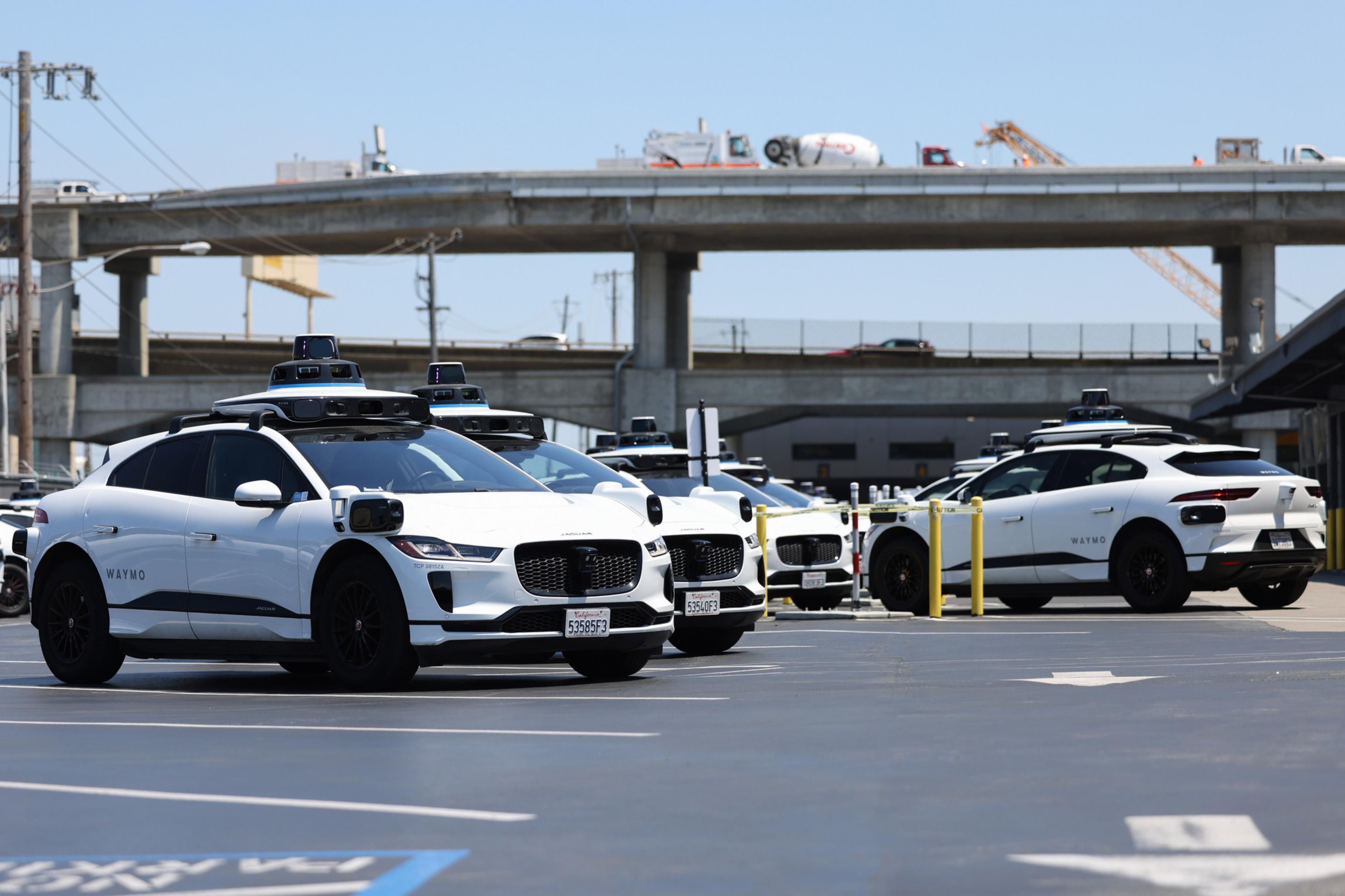 Three white autonomous vehicles with rooftop sensors are parked in a lot, with an overpass and cranes in the background.