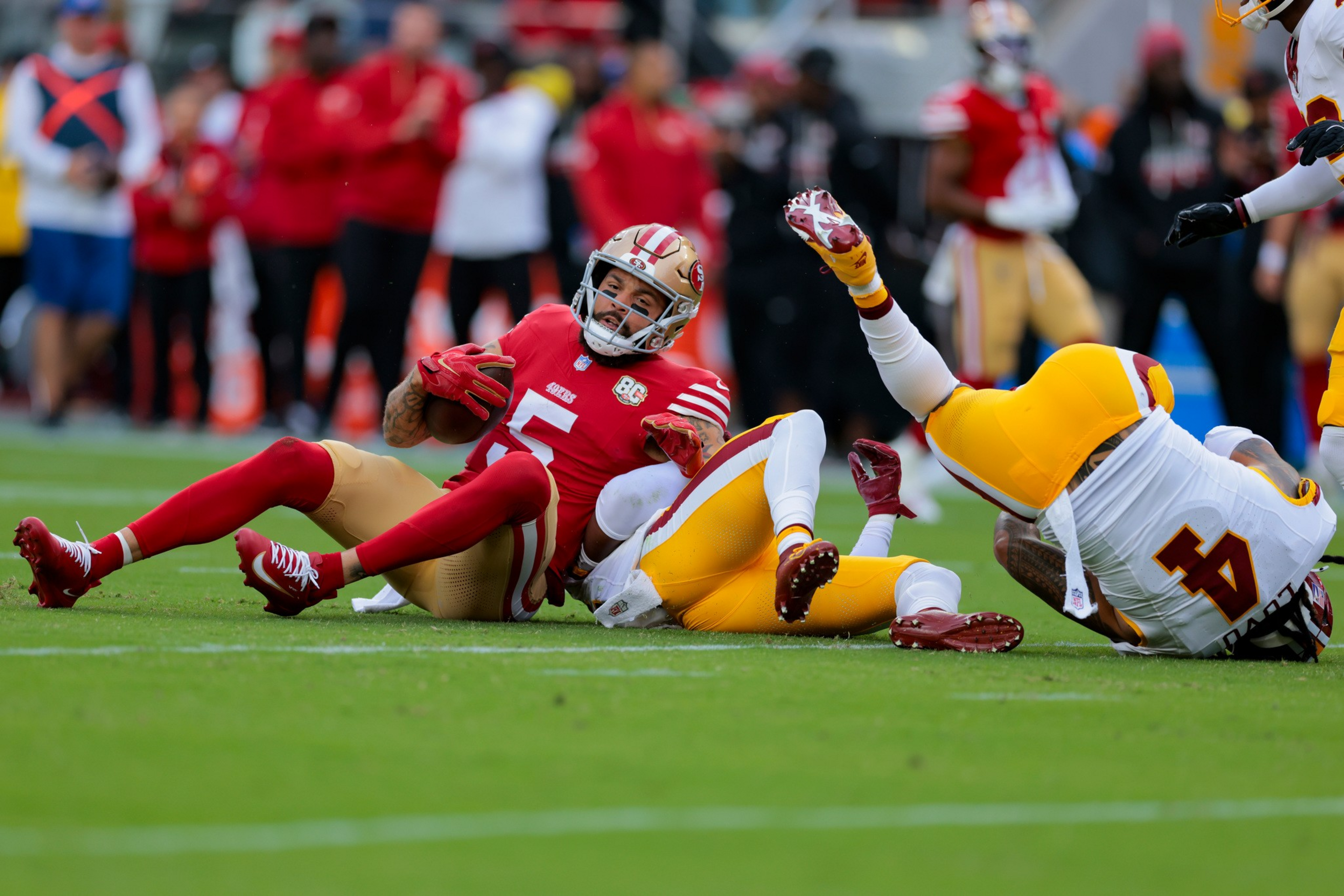 A football player in red and gold, holding the ball, is falling to the ground while two players in white and yellow tackle him.