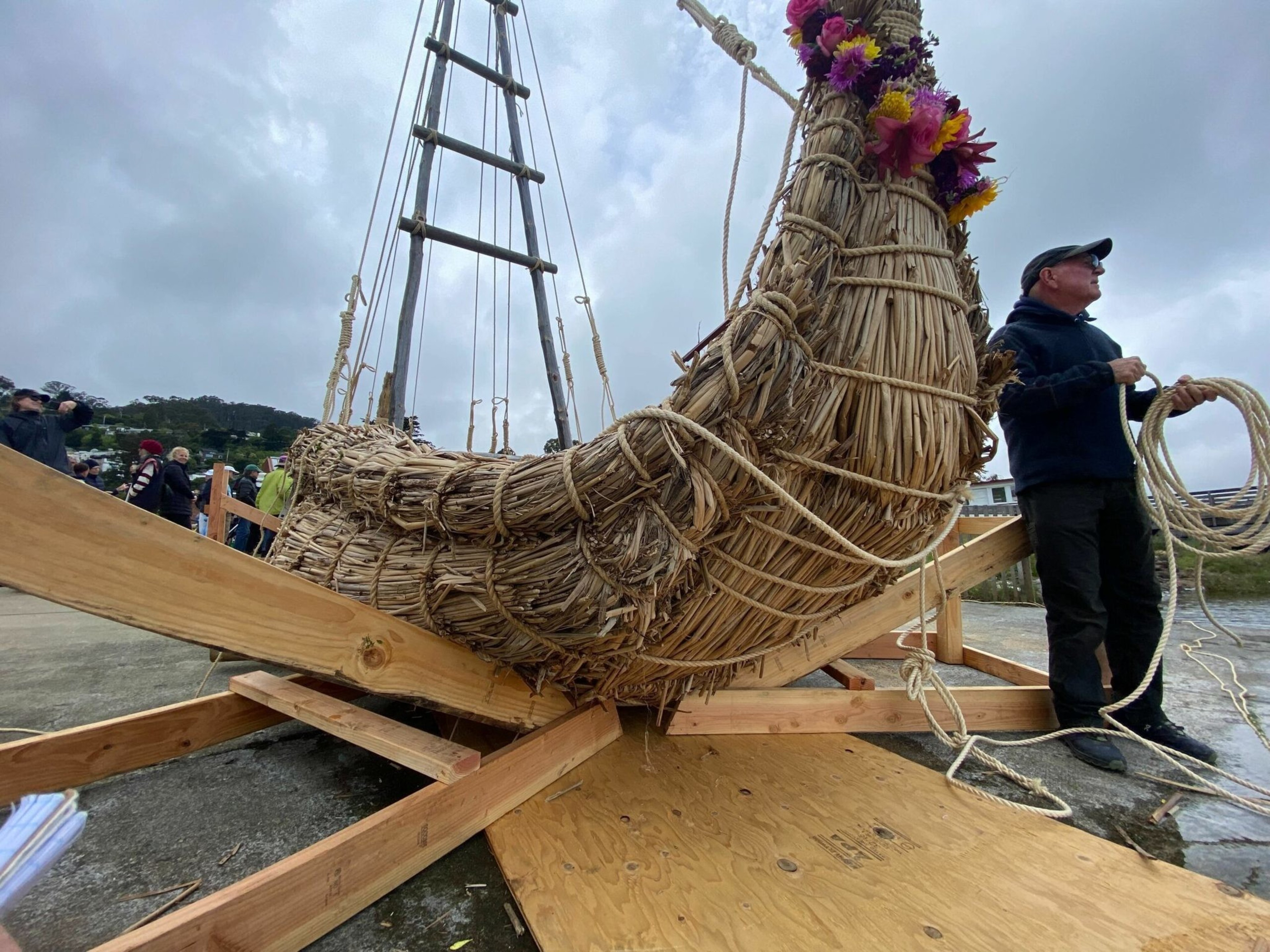 A large traditional reed boat decorated with flowers is supported by wooden beams, with a man holding coiled ropes standing nearby under a cloudy sky.