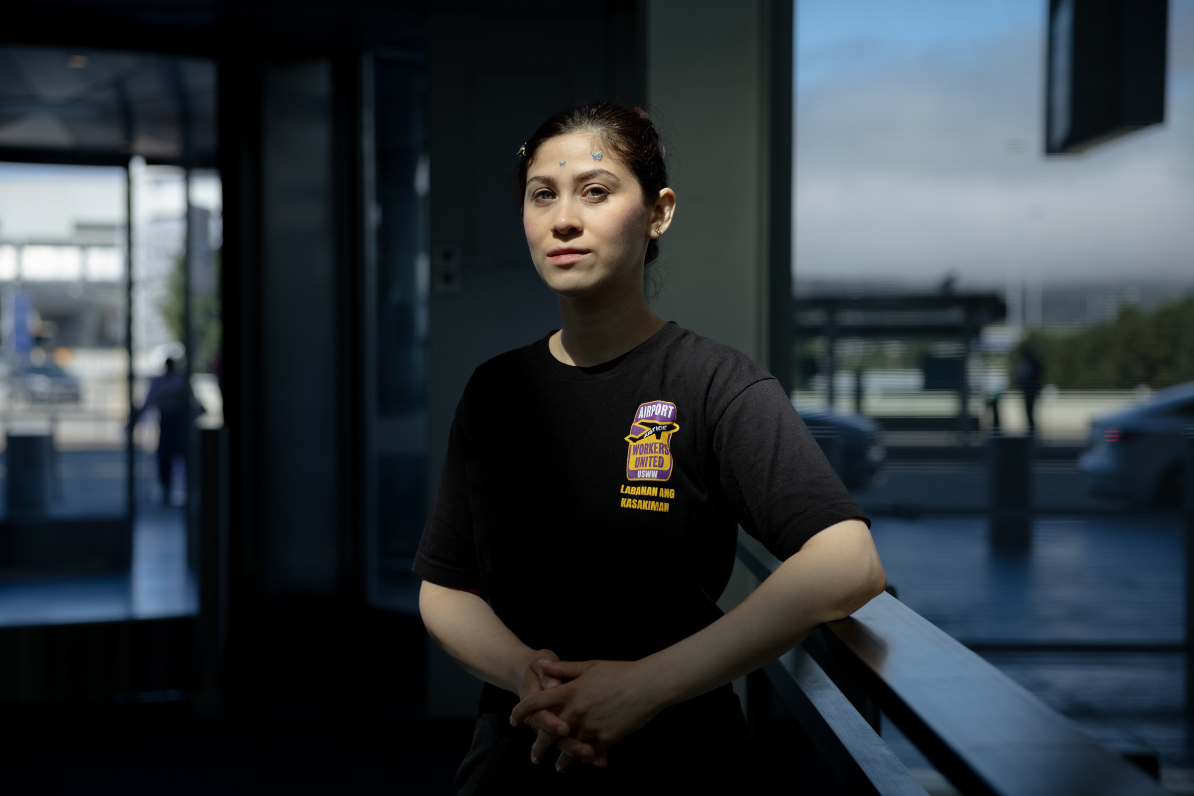 A woman with light skin and dark hair, wearing a black shirt with a colorful logo, stands indoors with her hands clasped, looking softly at the camera.