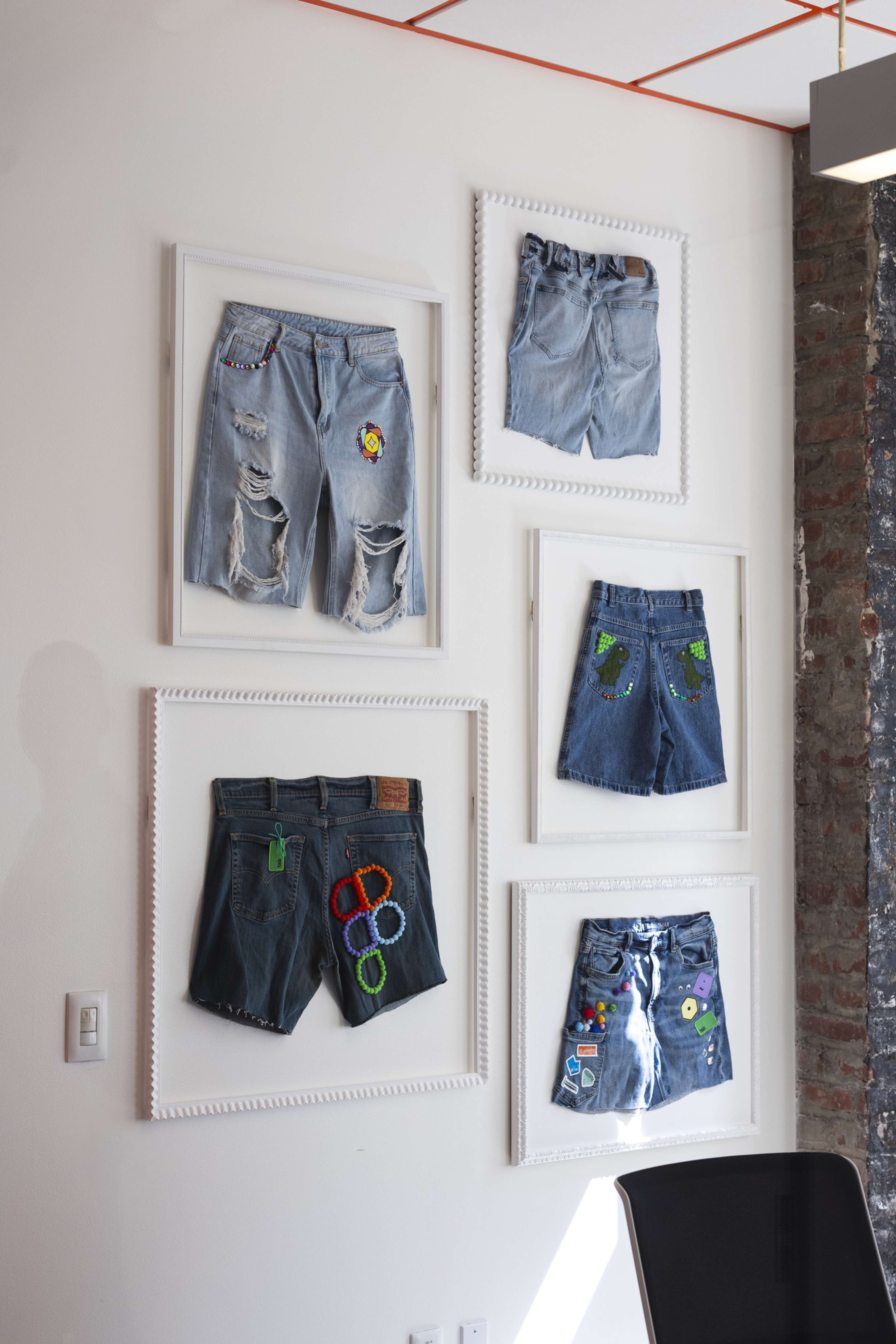 Five pairs of decorated denim shorts are framed and hung on a white wall, each featuring different patches, designs, and levels of distress.