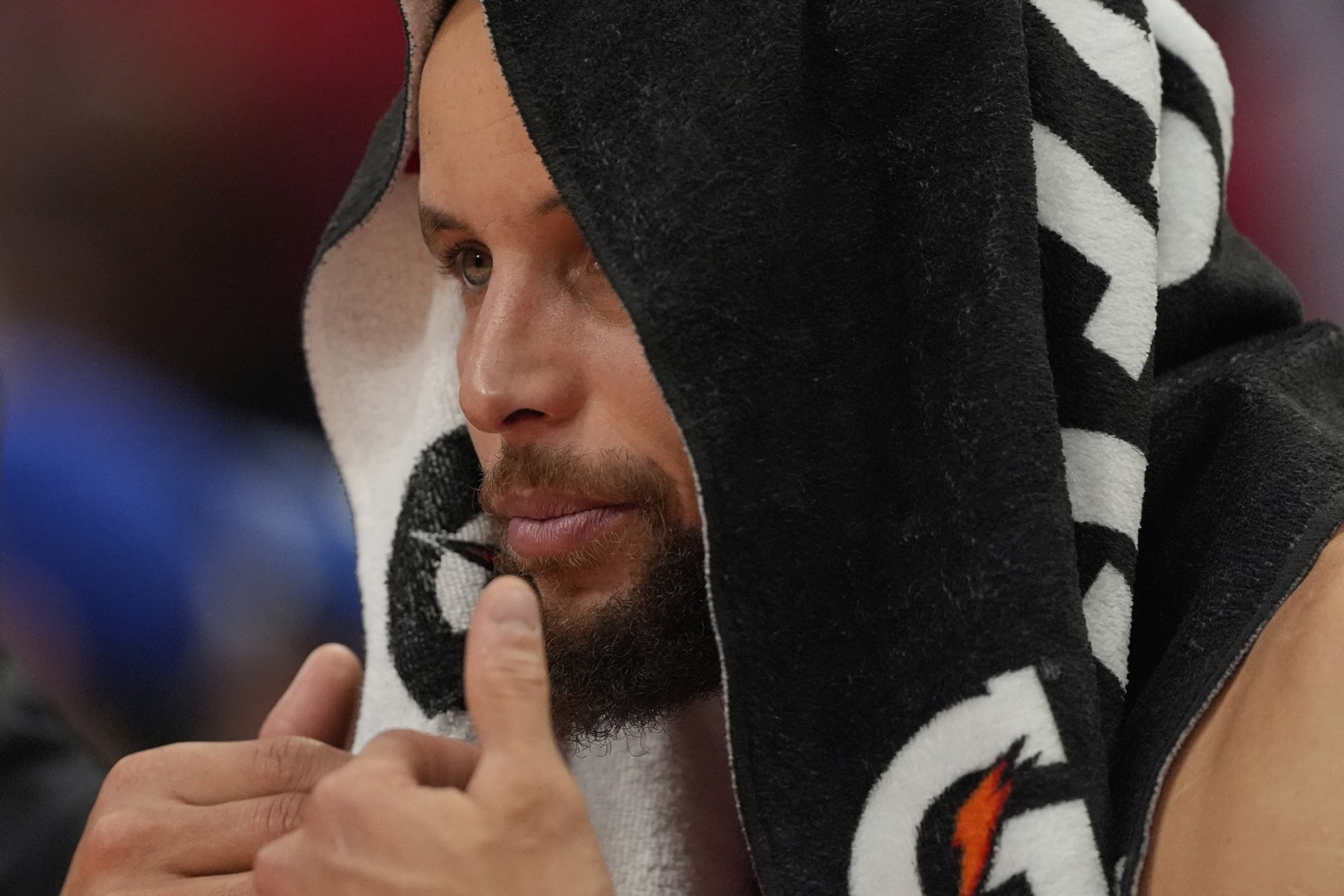A person is sitting with a towel draped over their head, displaying focus. The towel has a sports logo on it, and they appear to be in a sporting environment.
