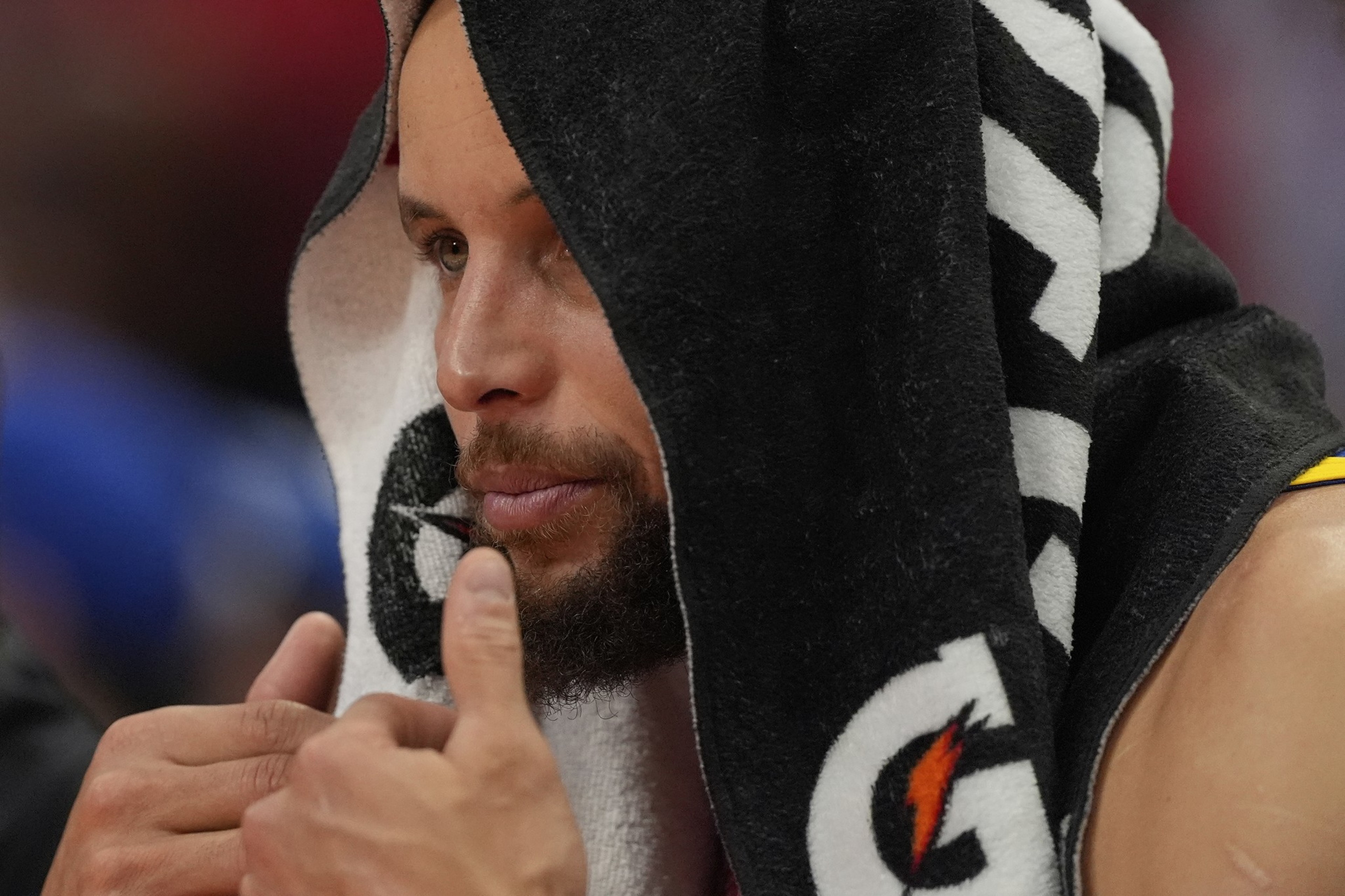 A person is sitting with a towel draped over their head, displaying focus. The towel has a sports logo on it, and they appear to be in a sporting environment.
