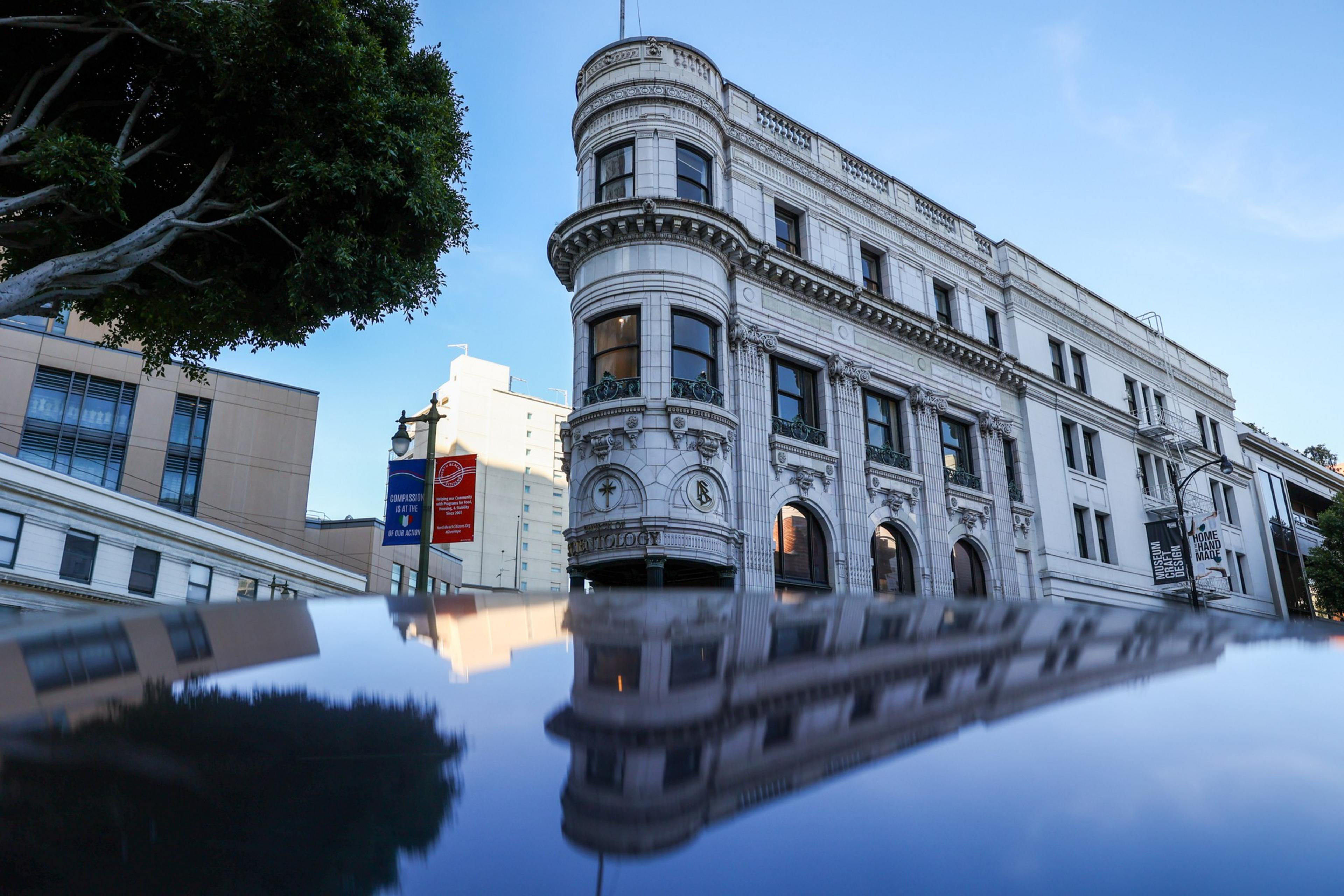 A historic white stone building with ornate details reflects clearly on a shiny black surface beneath a blue sky.