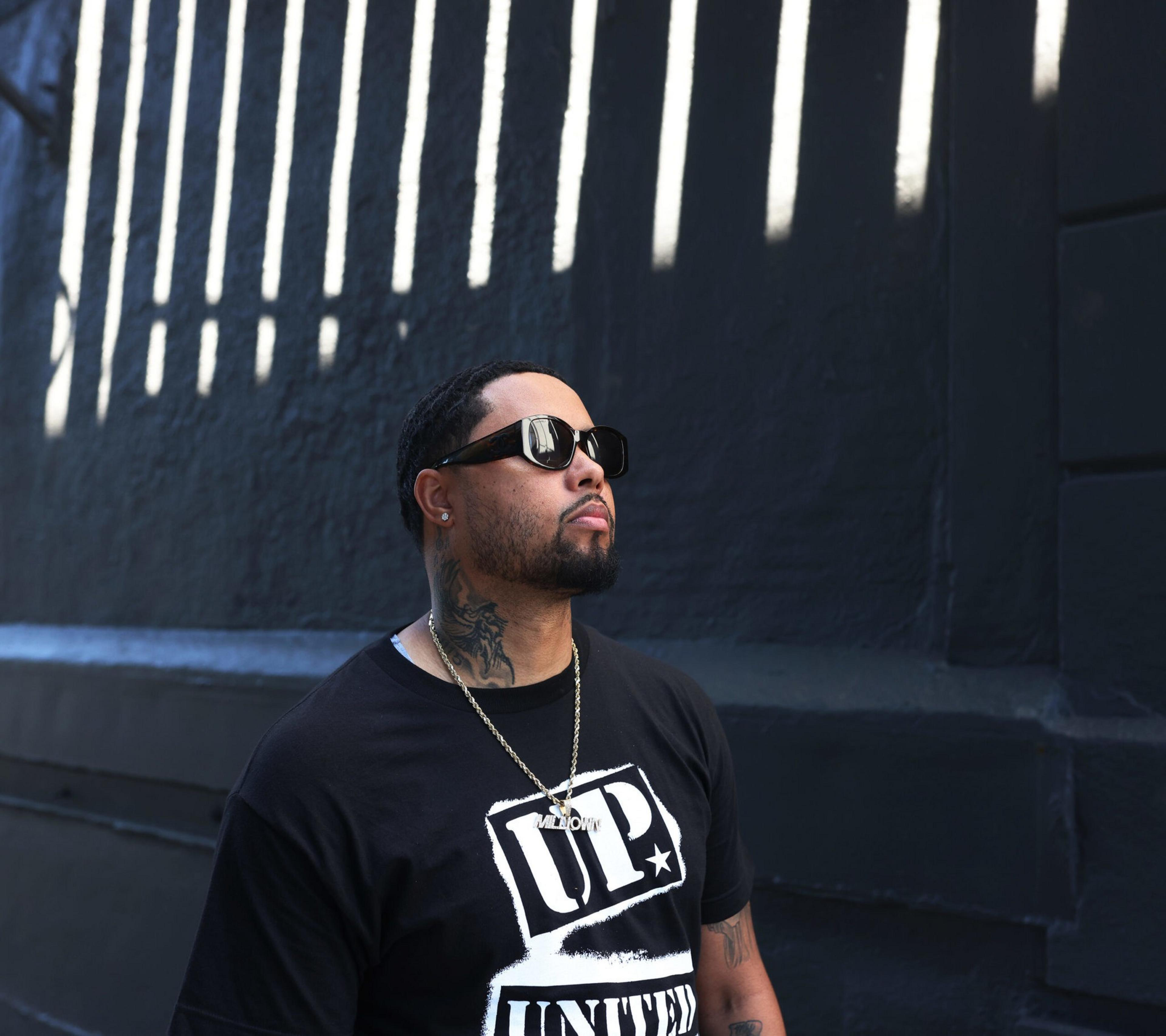 A man with braided hair, sunglasses, and a neck tattoo wears a black "UP UNITED" t-shirt and silver chain, standing against a dark wall with vertical light stripes.