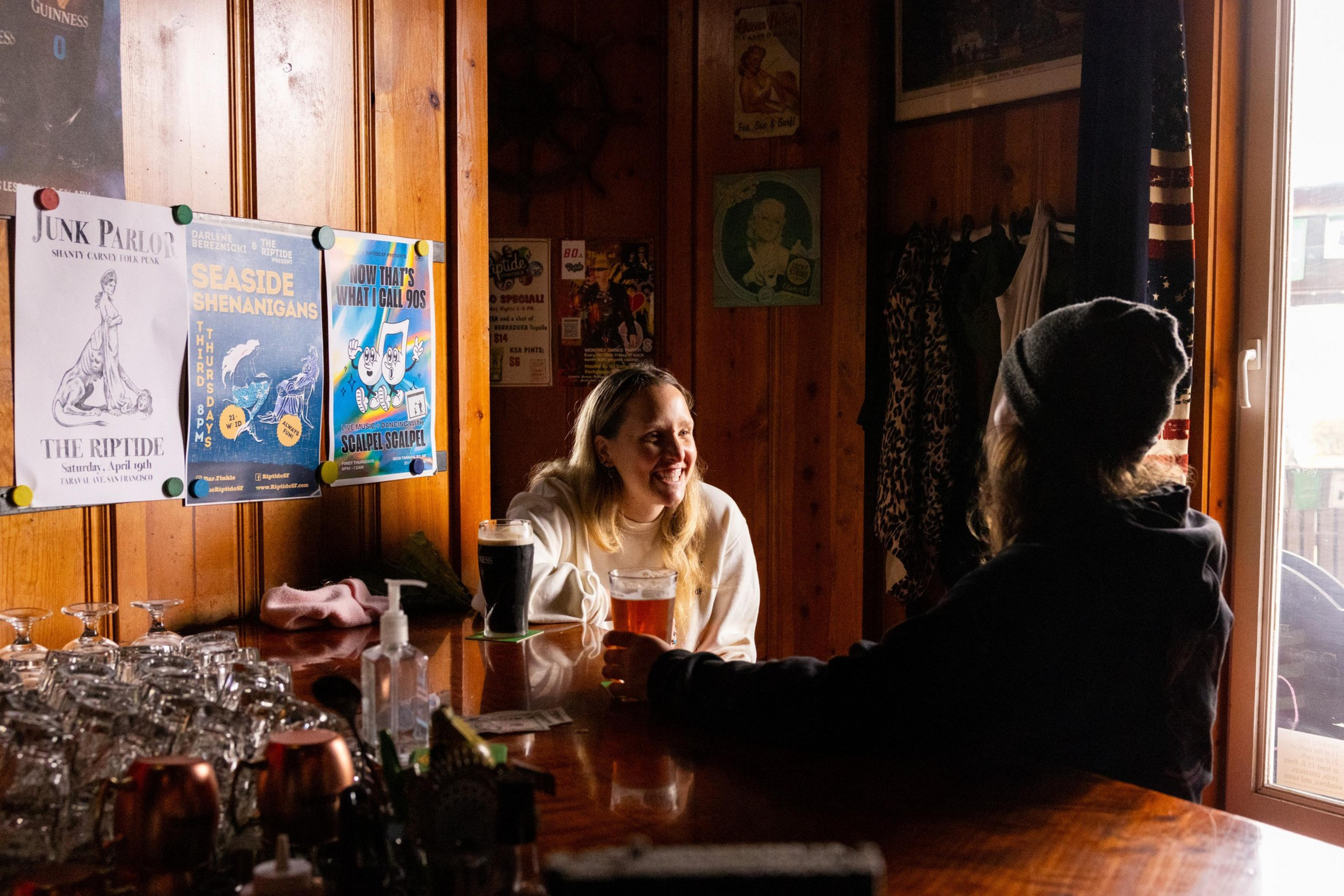 Two people sit at a wooden bar, engaged in conversation. One wears a beanie, and the other smiles warmly. Posters and glassware decorate the cozy setting.