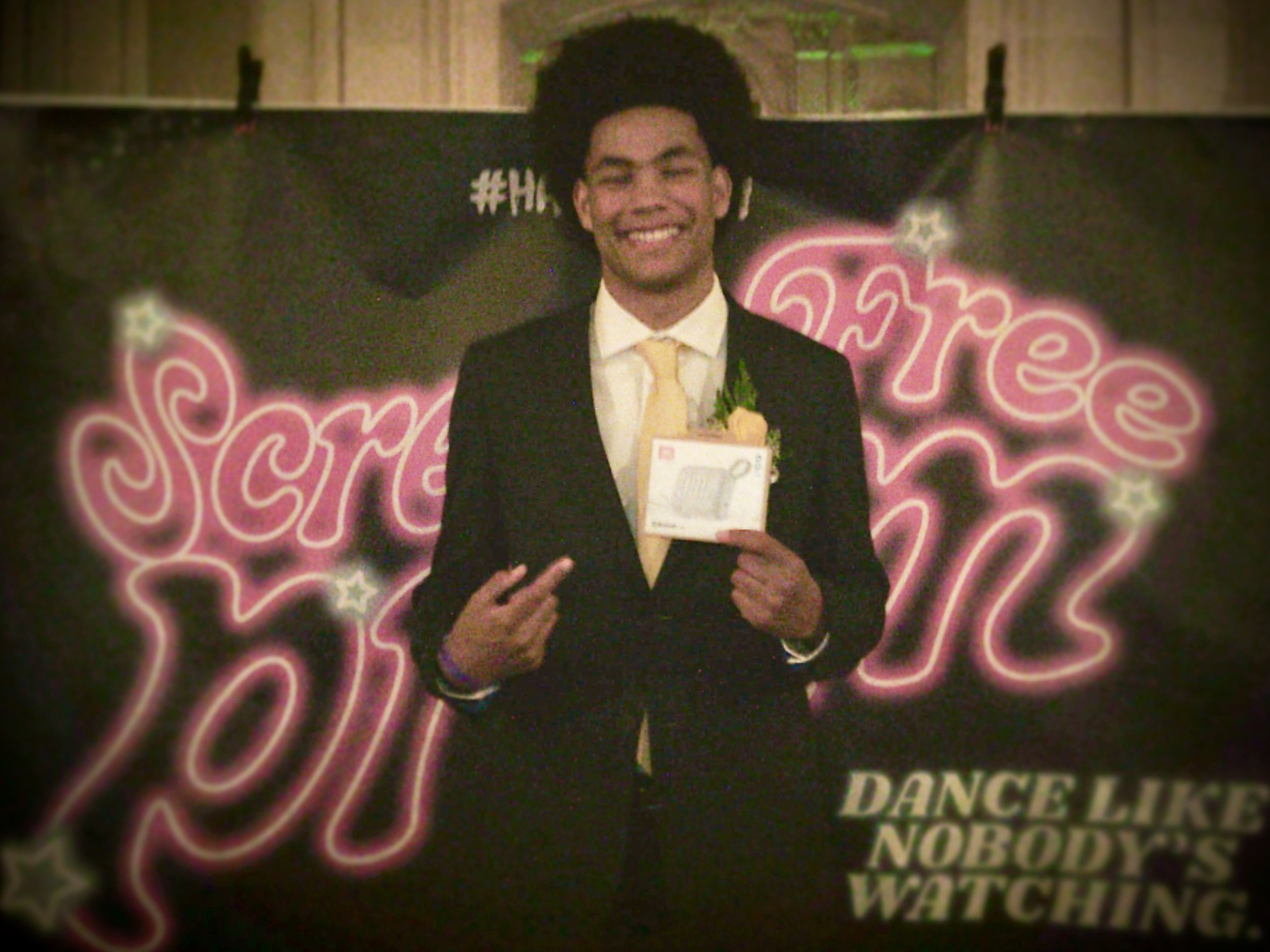 A person in a suit and yellow tie smiles, holding a small box. Behind them, a neon-lit backdrop says &quot;Scream Prom&quot; and &quot;Dance Like Nobody's Watching.&quot;