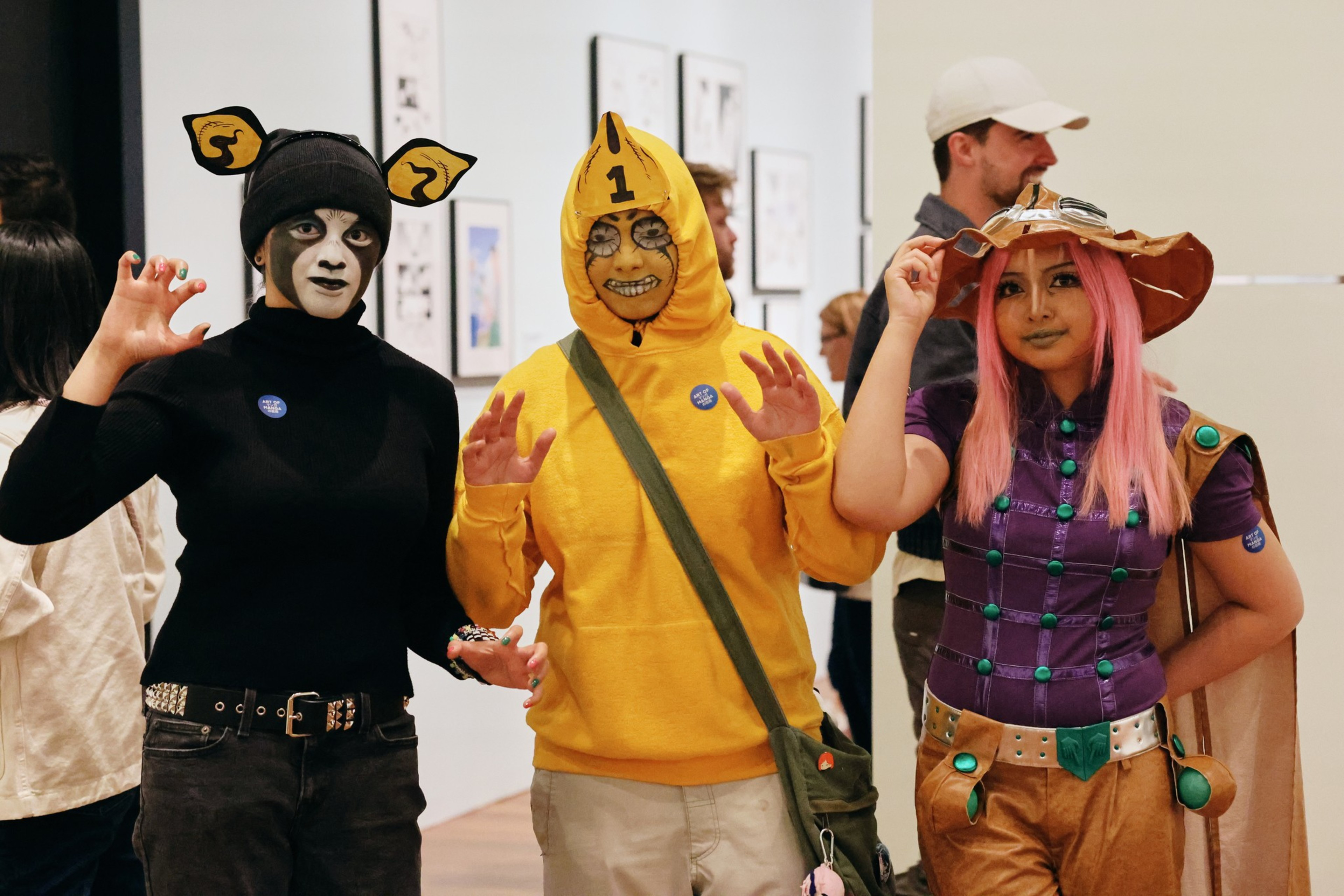 Three people in colorful costumes pose indoors; one wears black with animal face paint and ears, another in a yellow hoodie marked “1,” and the third in a purple outfit with a large hat.