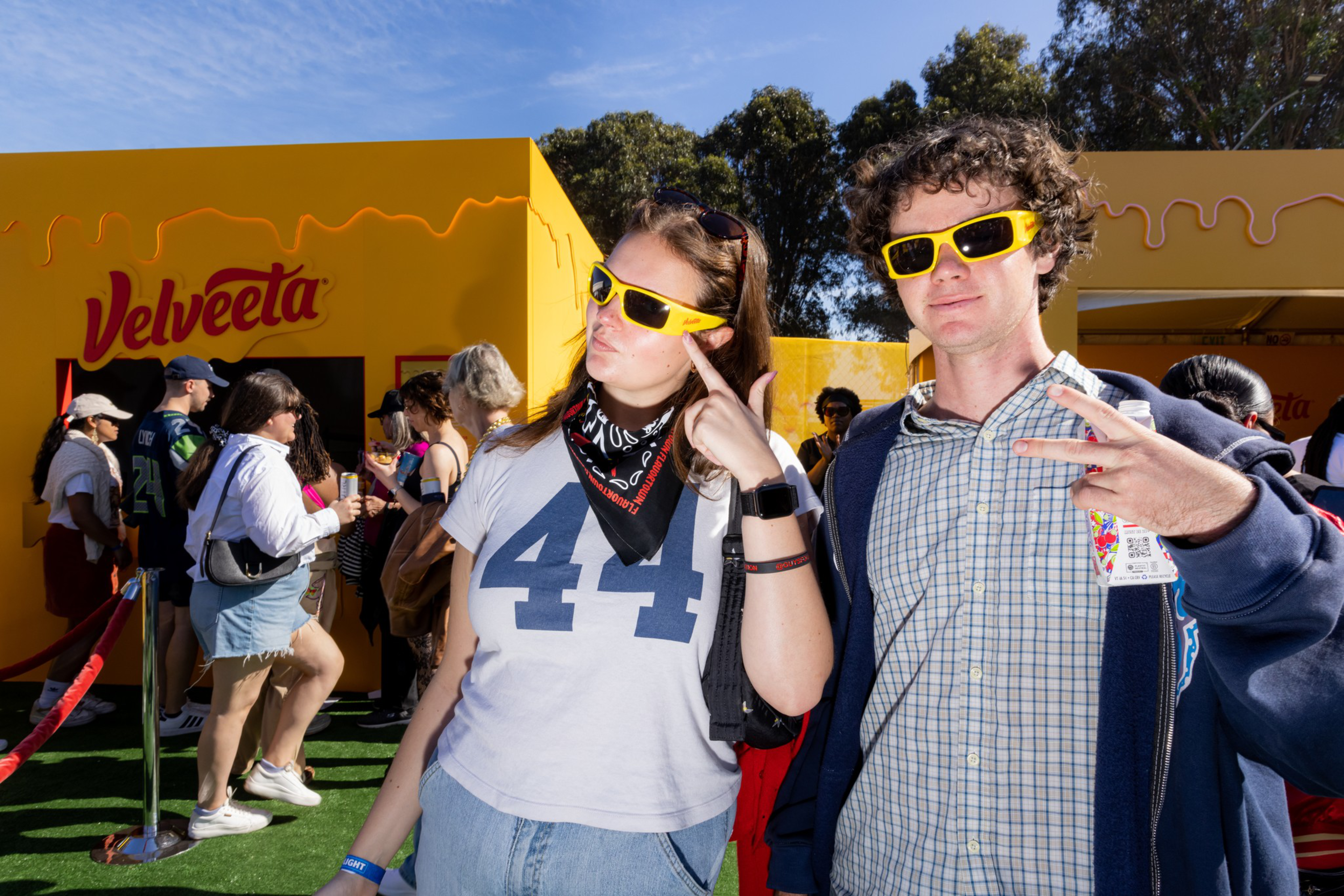 Two people wear yellow sunglasses and pose in front of a yellow Velveeta booth, while other people wait in line nearby on a sunny day.