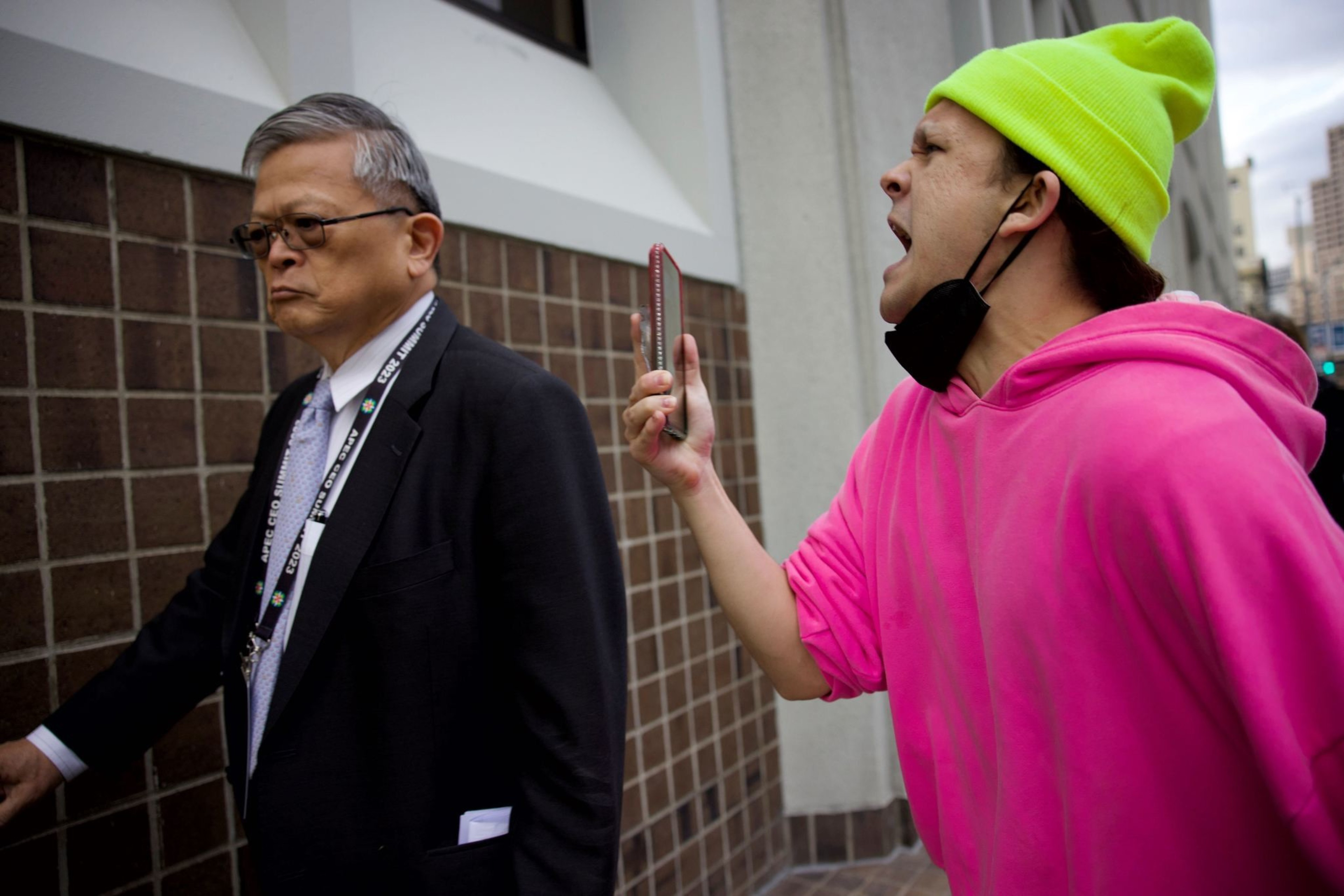 An angry person yells while holding a phone to record an APEC summit delegate as they walk along the sidewalk.