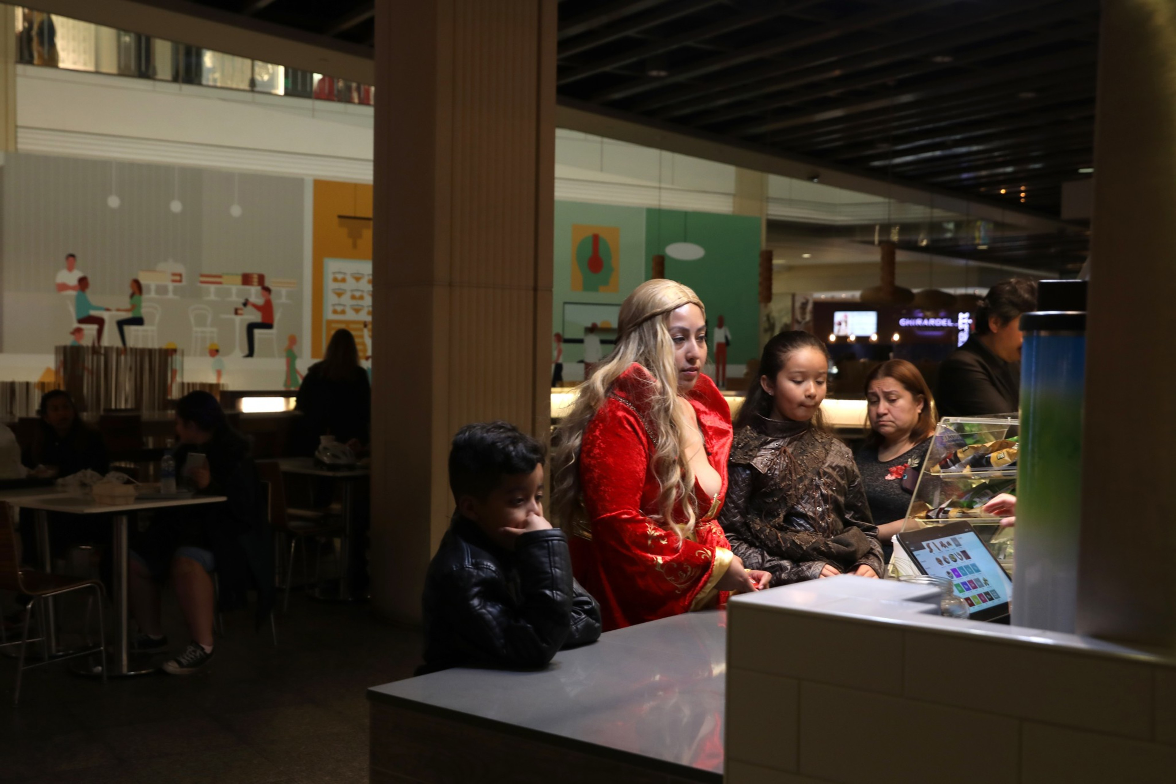 A family at a food counter, with a bored child leaning on his hand. People dine in the background.