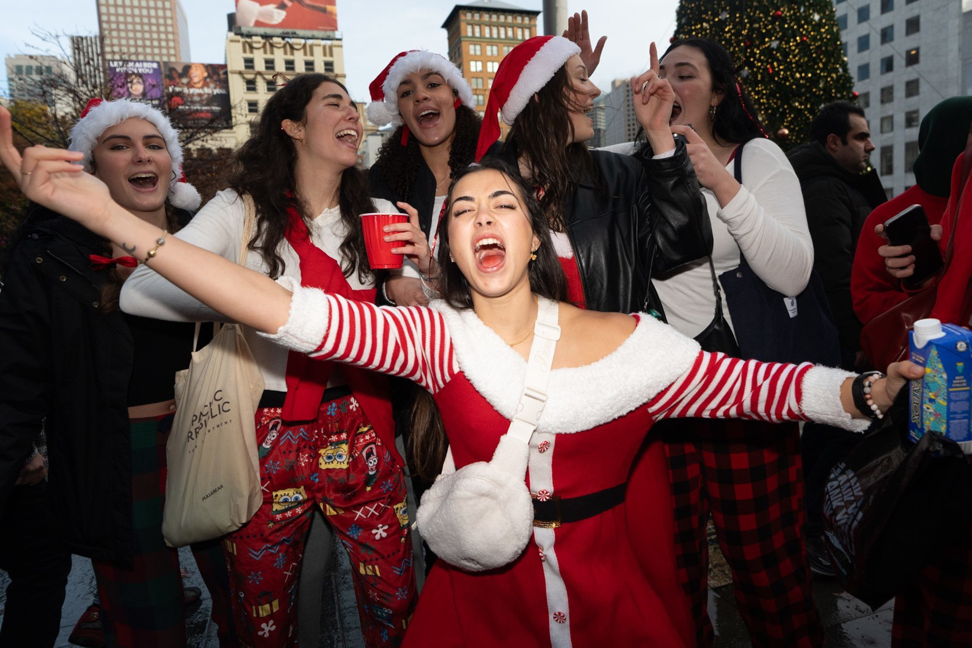 A joyful group of people, mostly women wearing festive Santa hats and red outfits, are celebrating outdoors near a decorated Christmas tree.