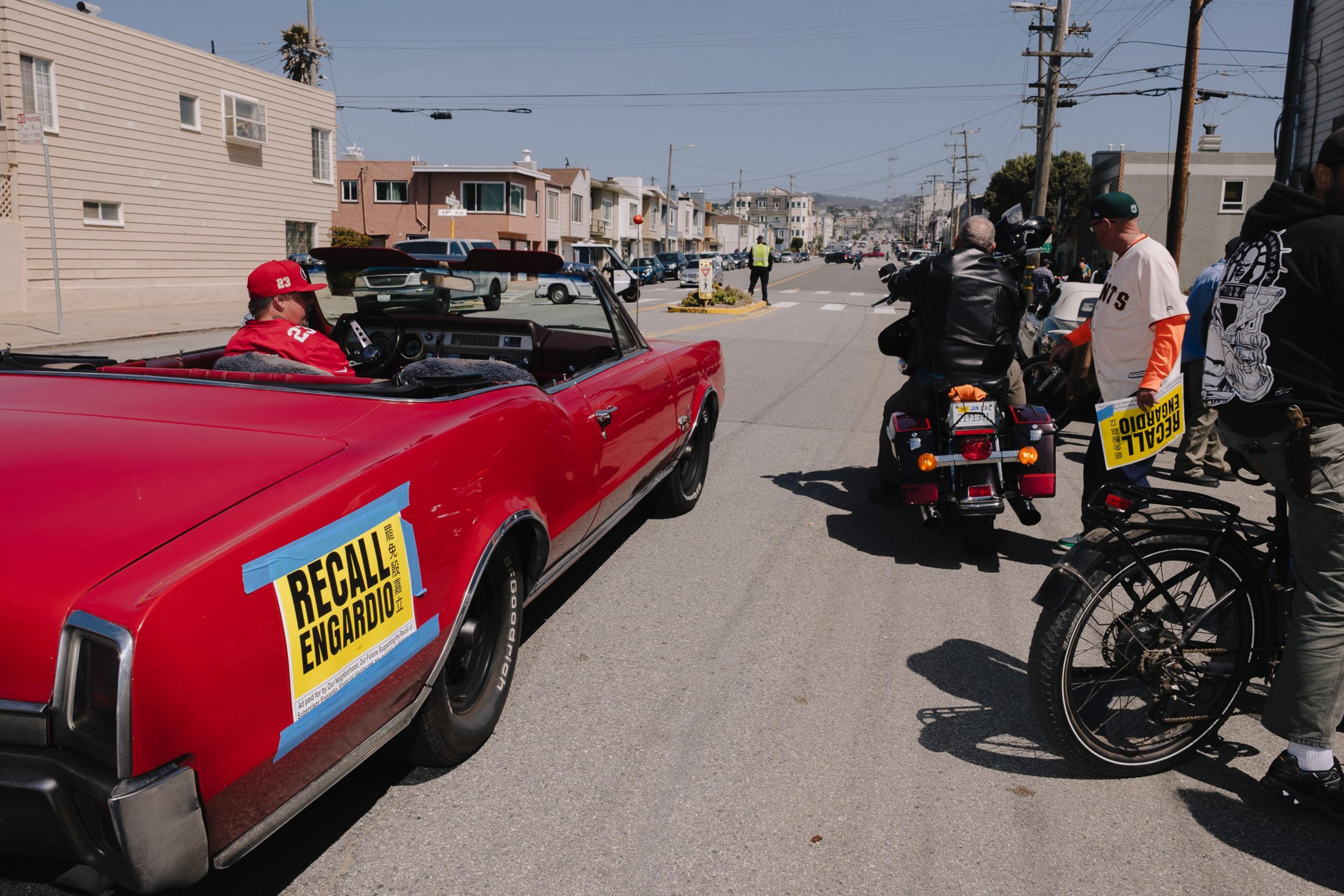 A red convertible with a &quot;Recall Engardio&quot; sign is in the street alongside a motorcycle and bicycle. Nearby people also hold &quot;Recall&quot; signs.