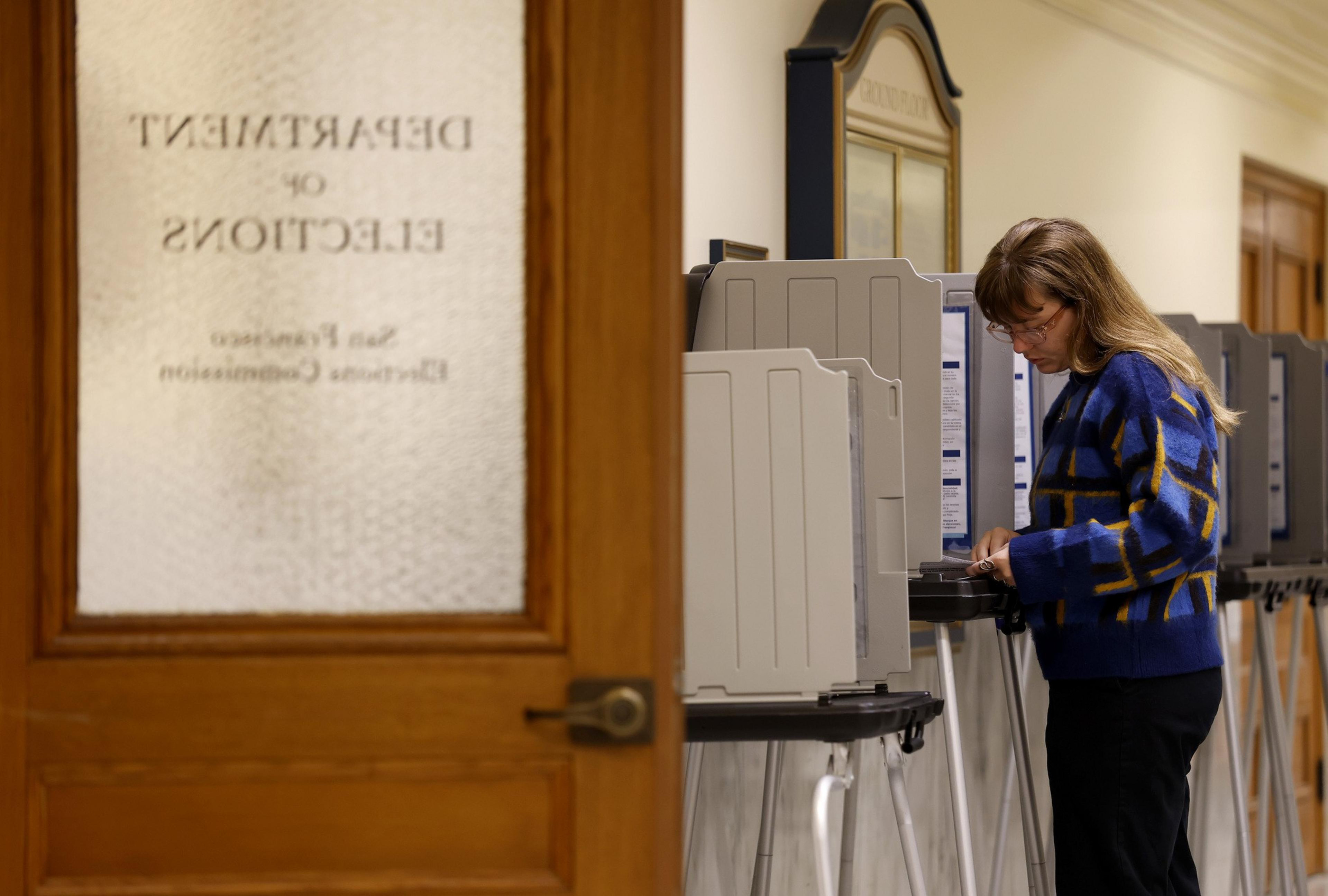 A woman wearing a blue patterned sweater stands at a voting booth inside a building, next to a door labeled “Department of Elections.”