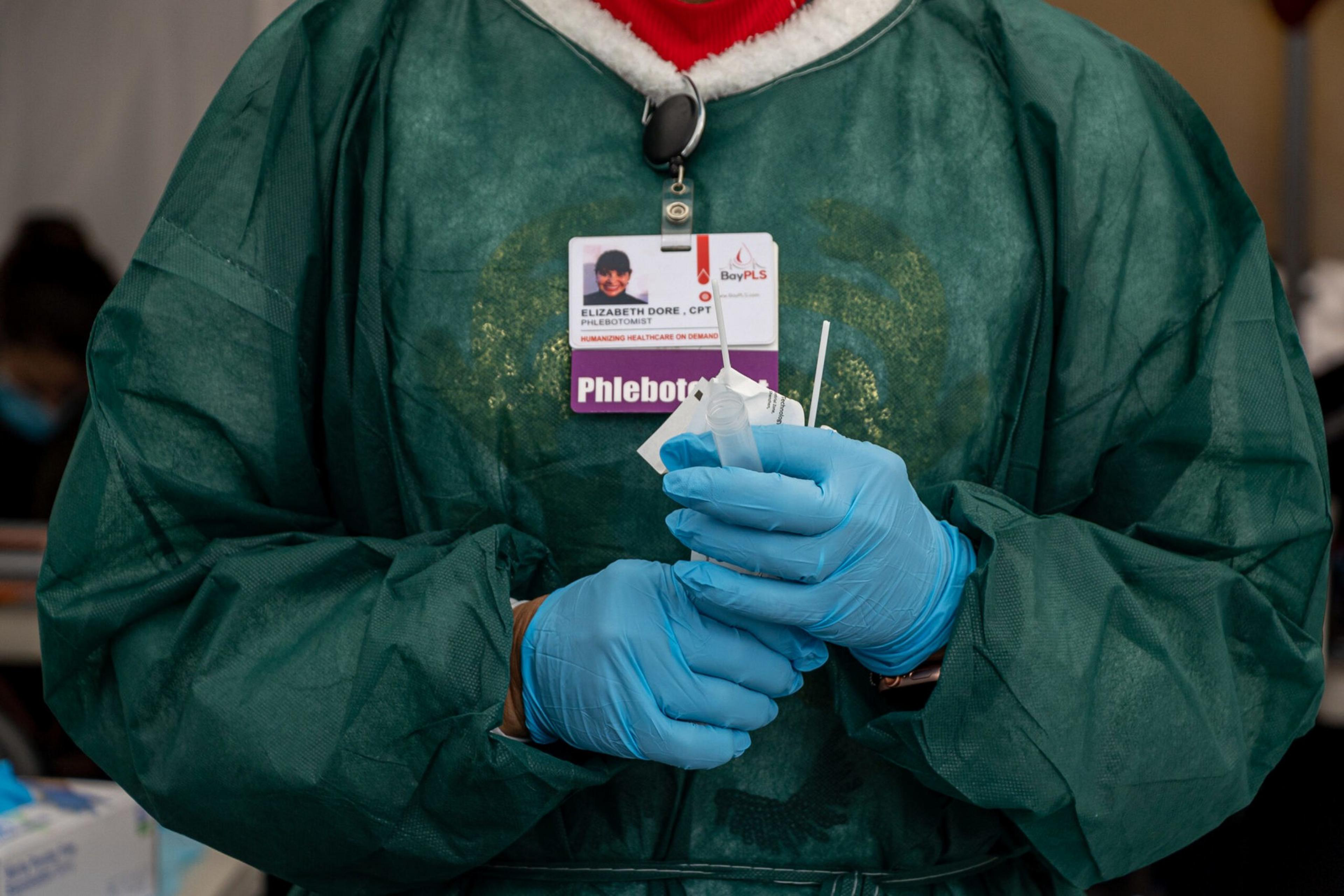 A person wearing blue gloves and a green protective gown holds medical swabs and testing materials, with a badge labeled "Phlebotomist."