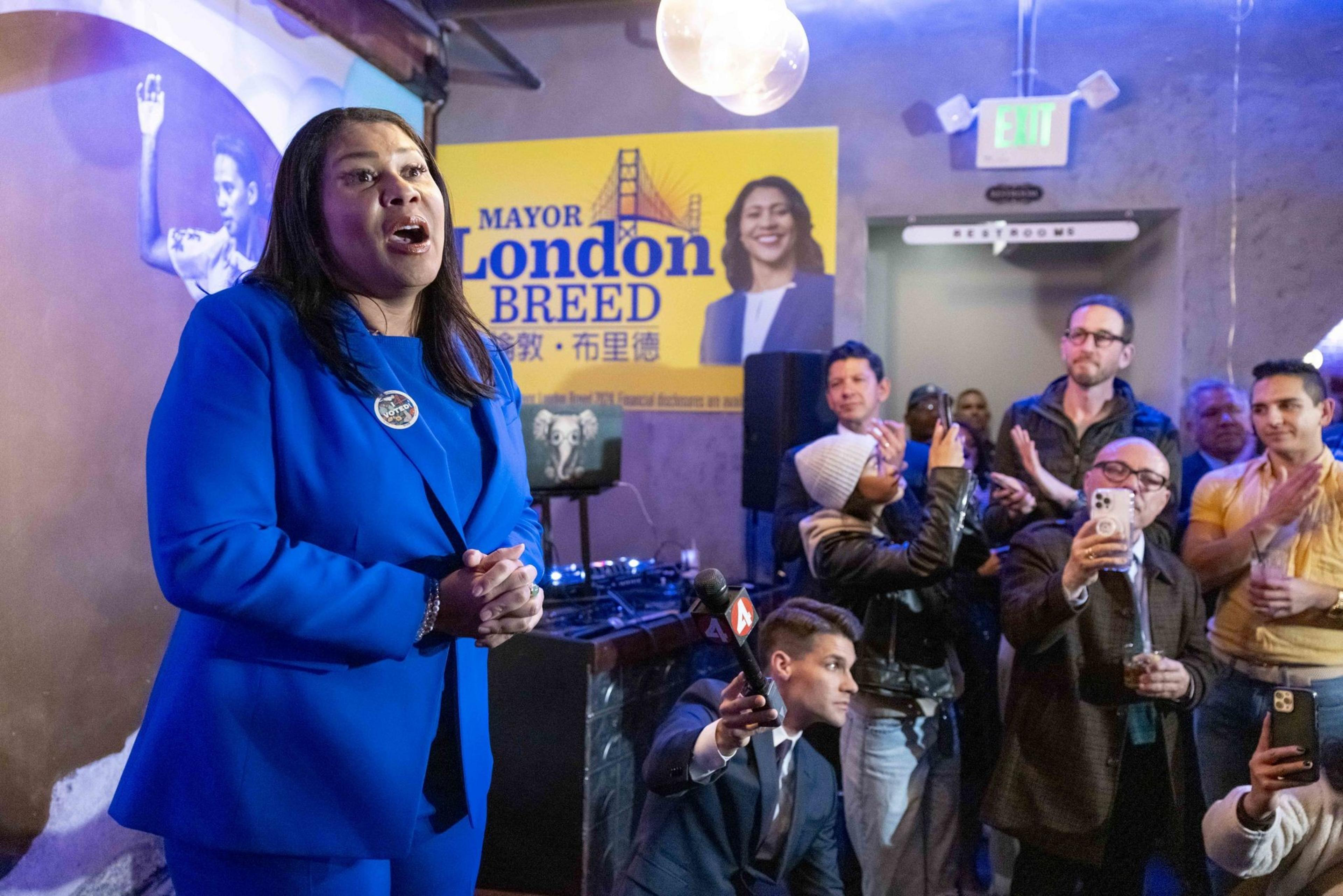 A woman in a blue suit speaks to a crowd in a room with a banner featuring her name. People are taking photos and recording with their phones.
