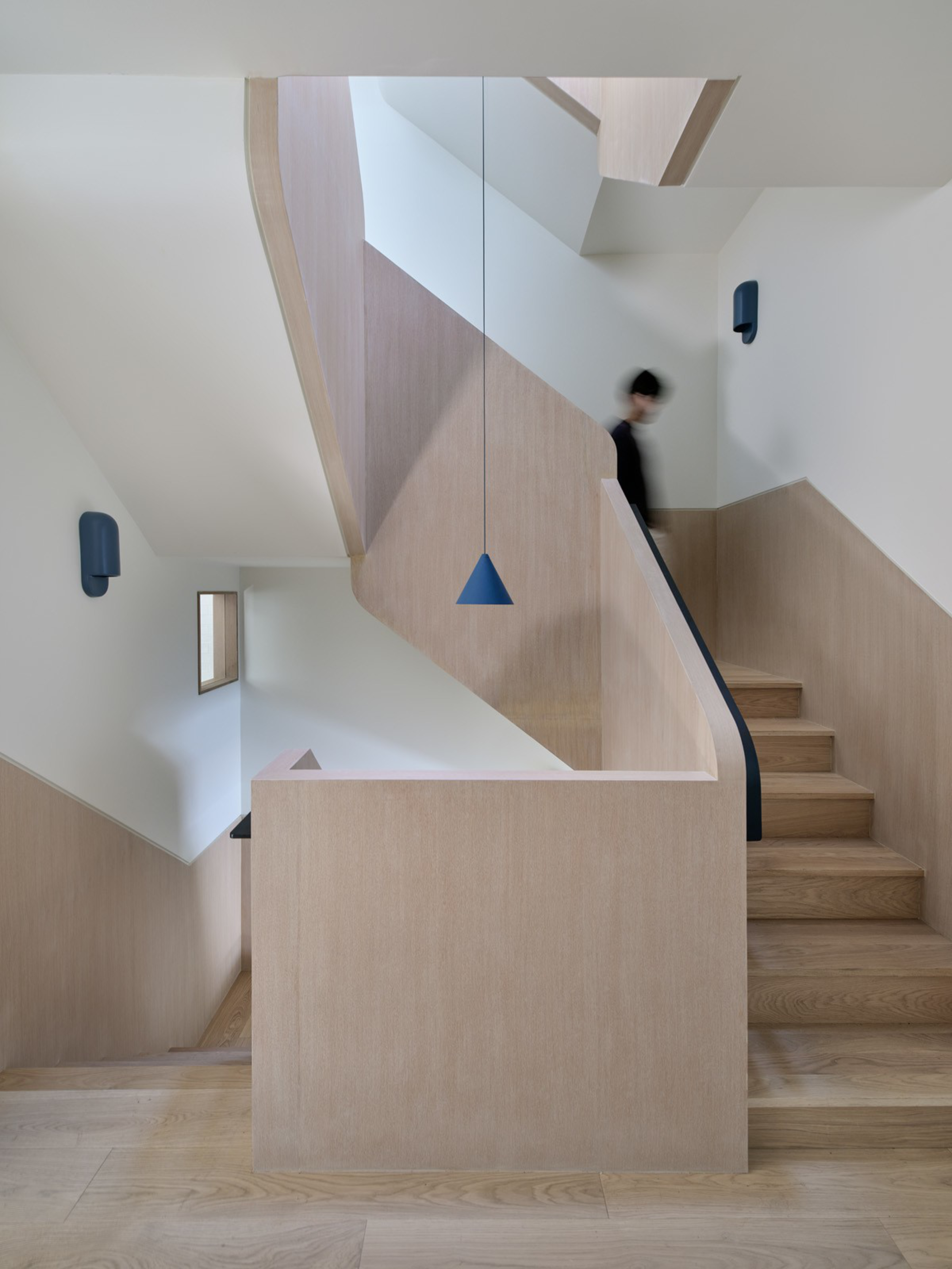 A modern wooden staircase with light wood paneling, a blue pendant light, and blurred person walking down steps in a minimal white-walled interior.