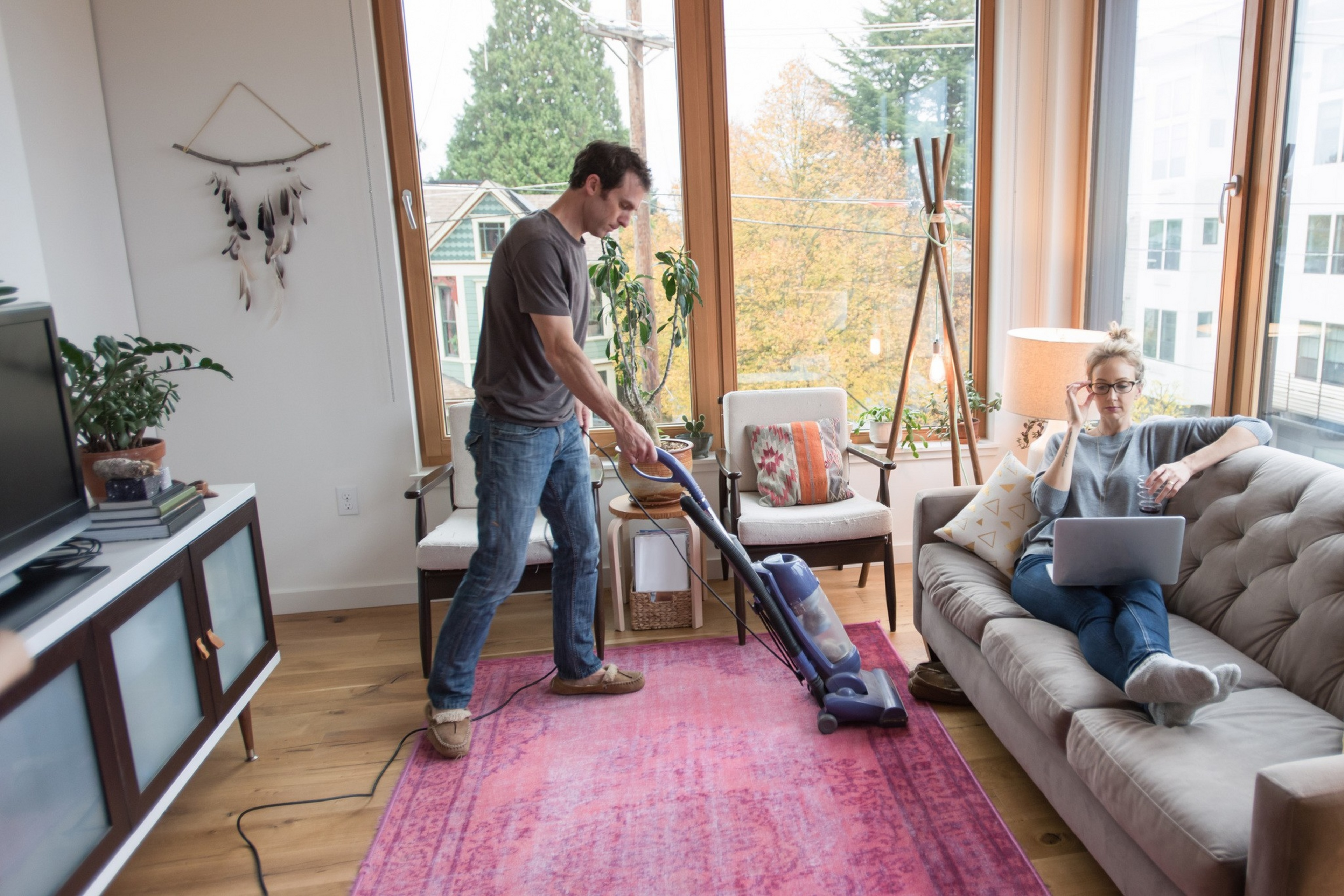 A man vacuuming a pink rug while a woman sits on a gray sofa using a laptop, in a cozy living room with large windows and plants.