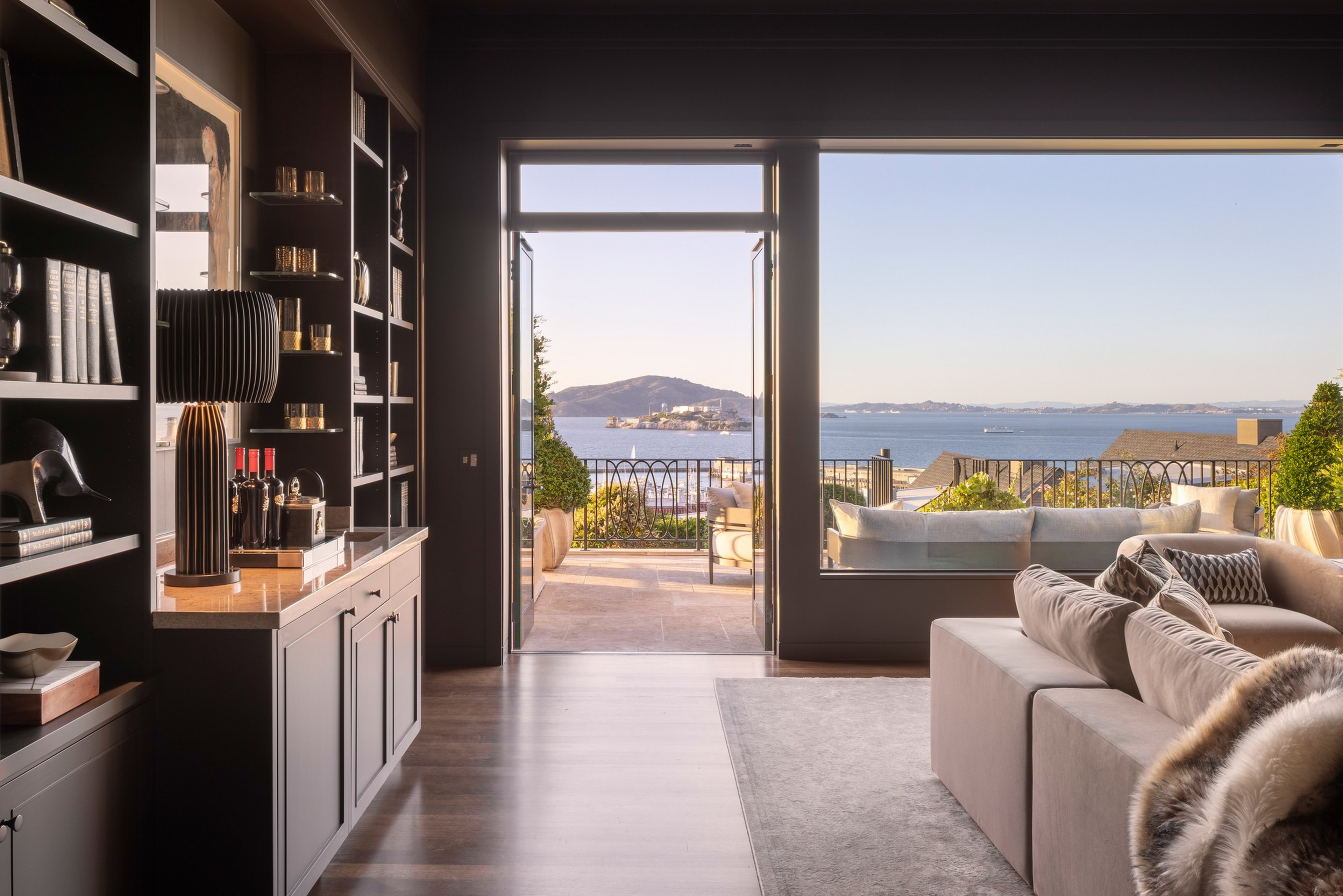 A modern living room with a beige sofa and built-in shelves opens to a terrace overlooking a large body of water and distant hills under a clear sky.