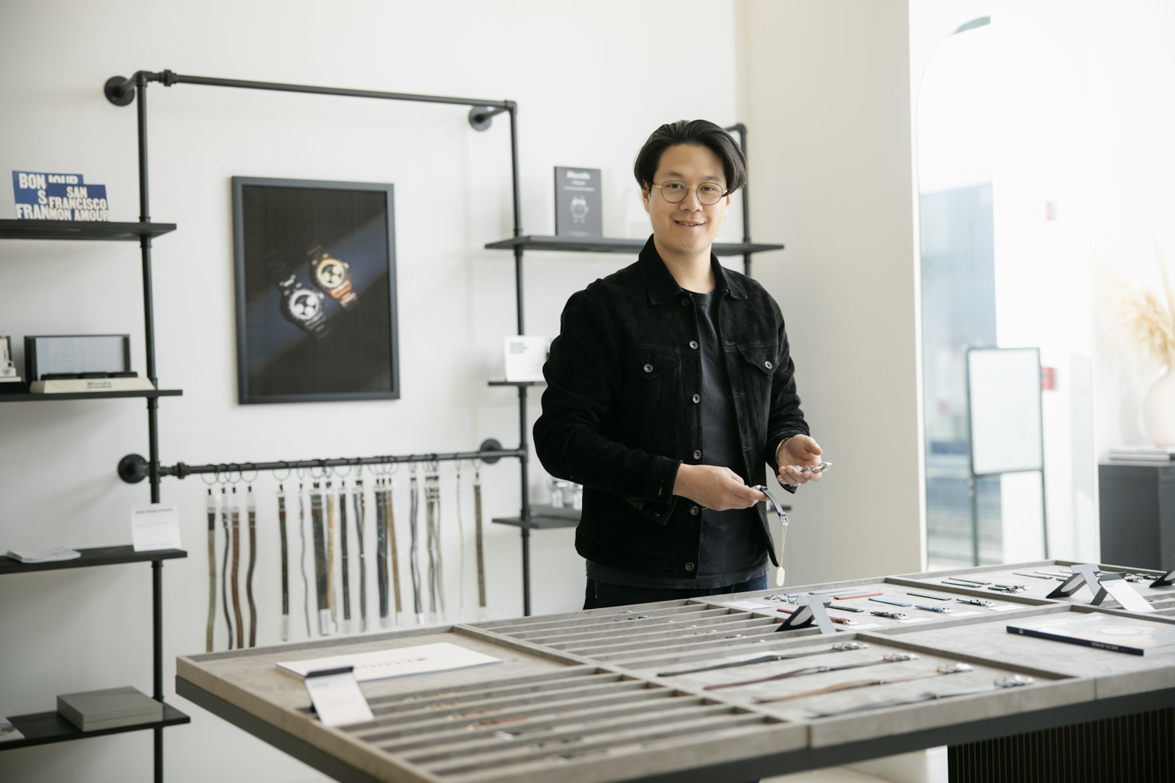 A man wearing glasses and a black jacket stands behind a display table in a watch store, holding a wristwatch in his hands.
