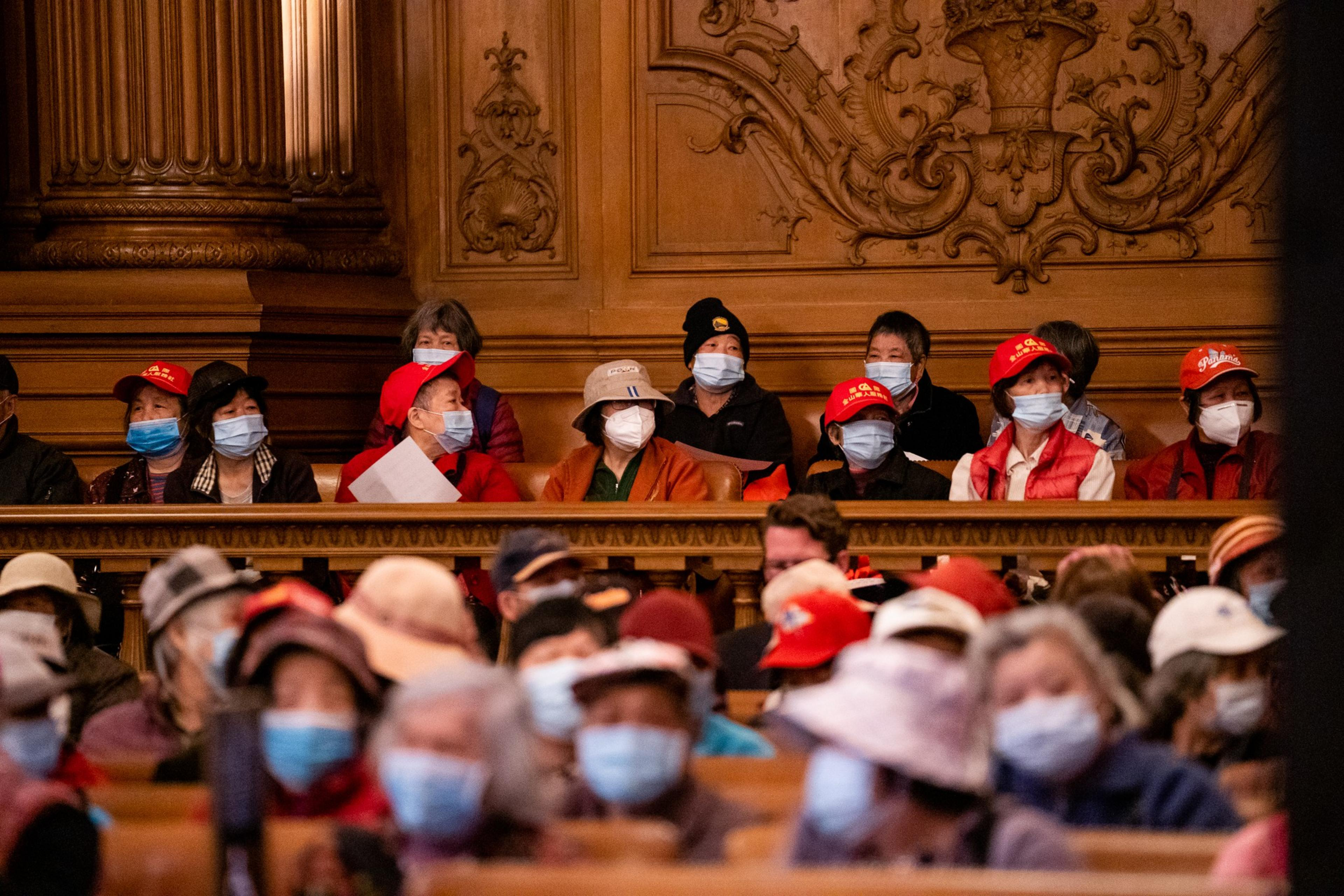 A group of people wearing face masks and hats sit in a wooden, ornately carved auditorium, with some wearing red caps and jackets.